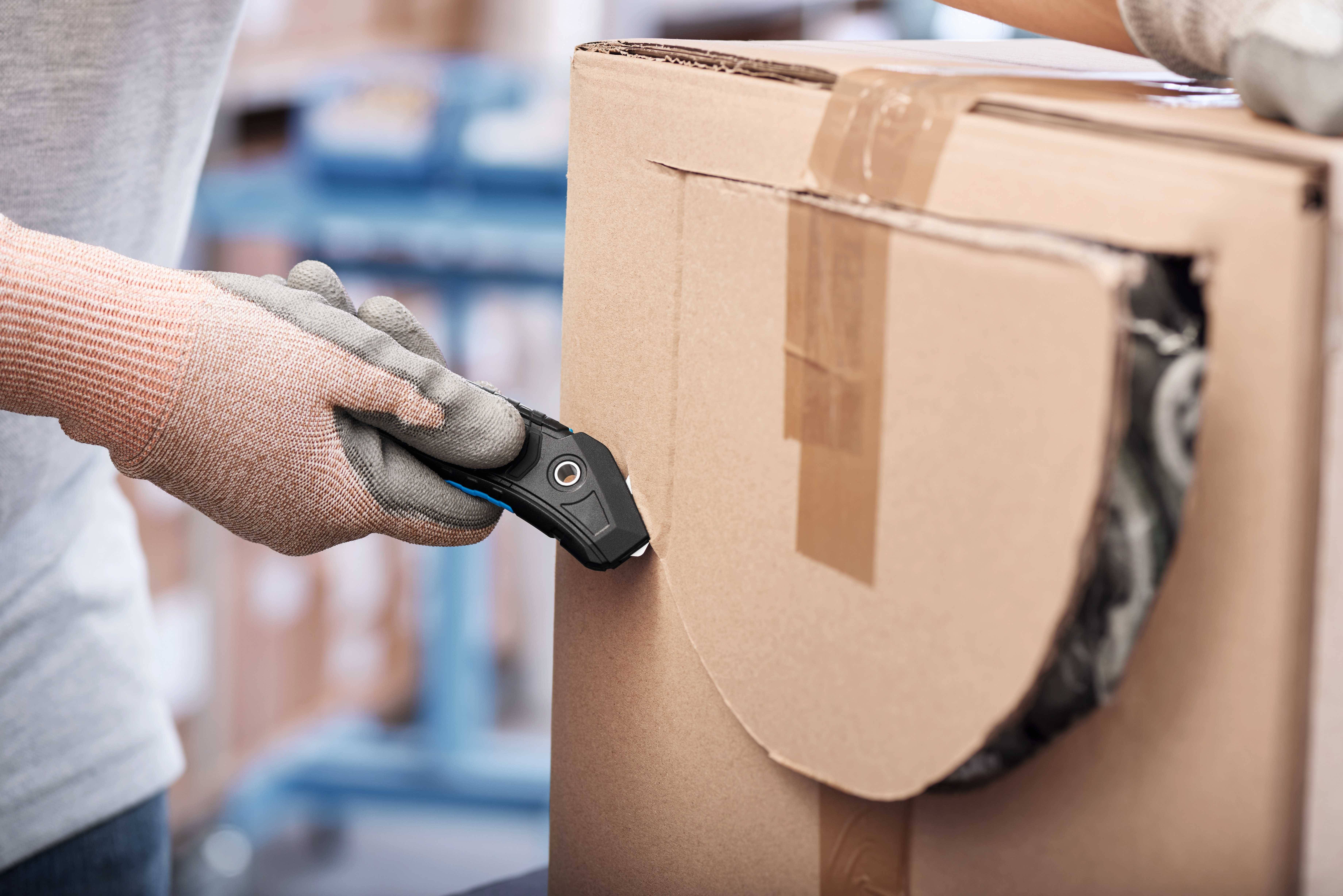 En person med arbejdshandsker skærer et stort papkasse op med en sikkerhedskniv.
