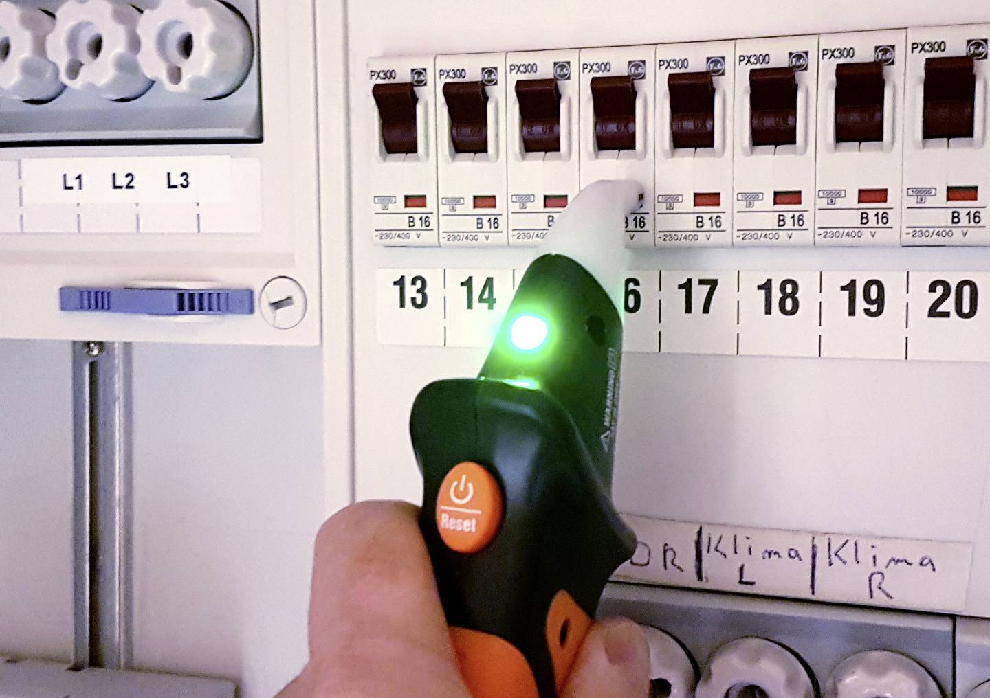 En hånd holder en spændingstester med et lysende grønt lys ved en sikringstavle for at verificere, at der ikke er spænding.