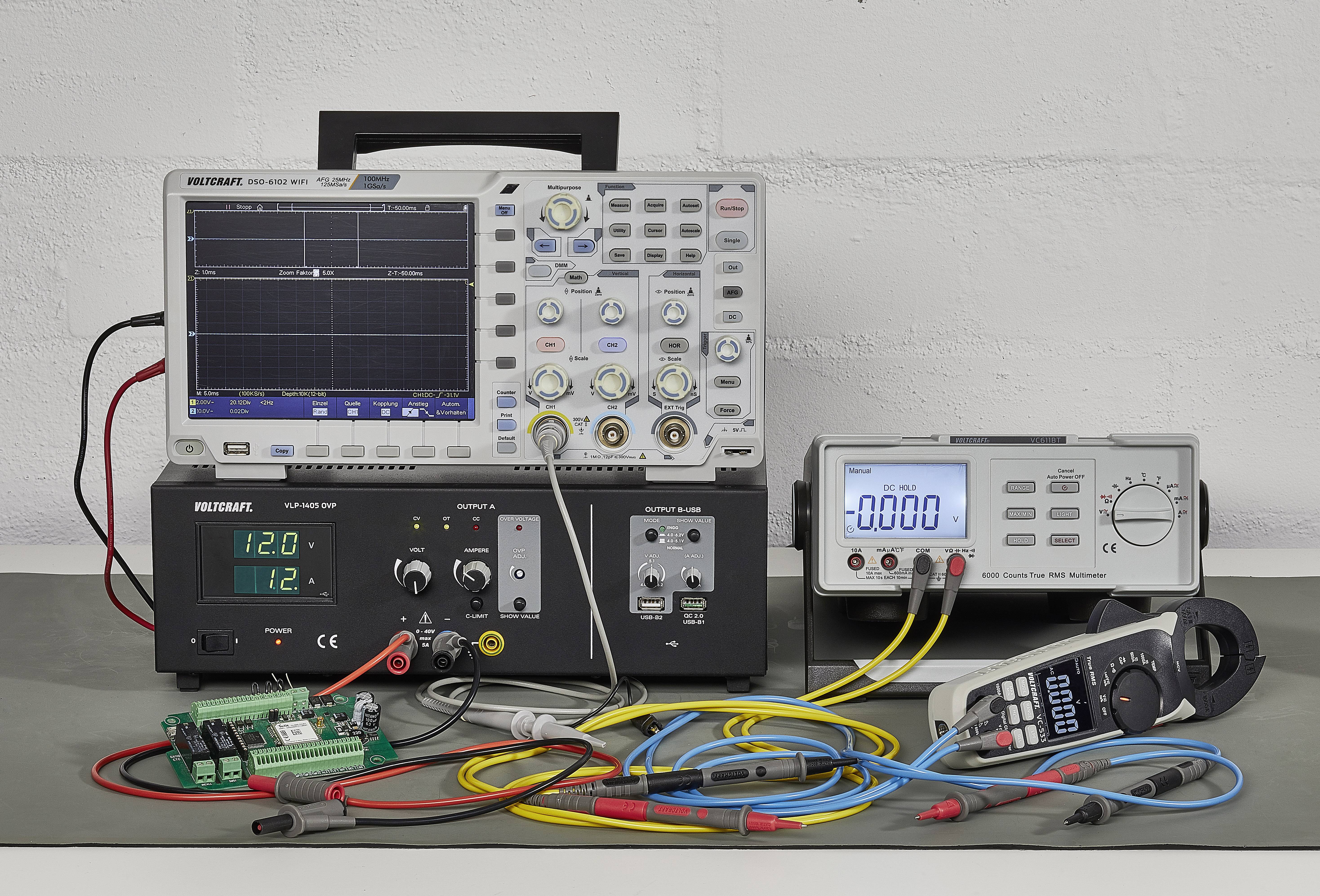 Et oscilloskop med tilsluttede kabler viser en kurve. Ved siden af ligger et multimeter og en strømforsyning, der viser digitale måleværdier.
