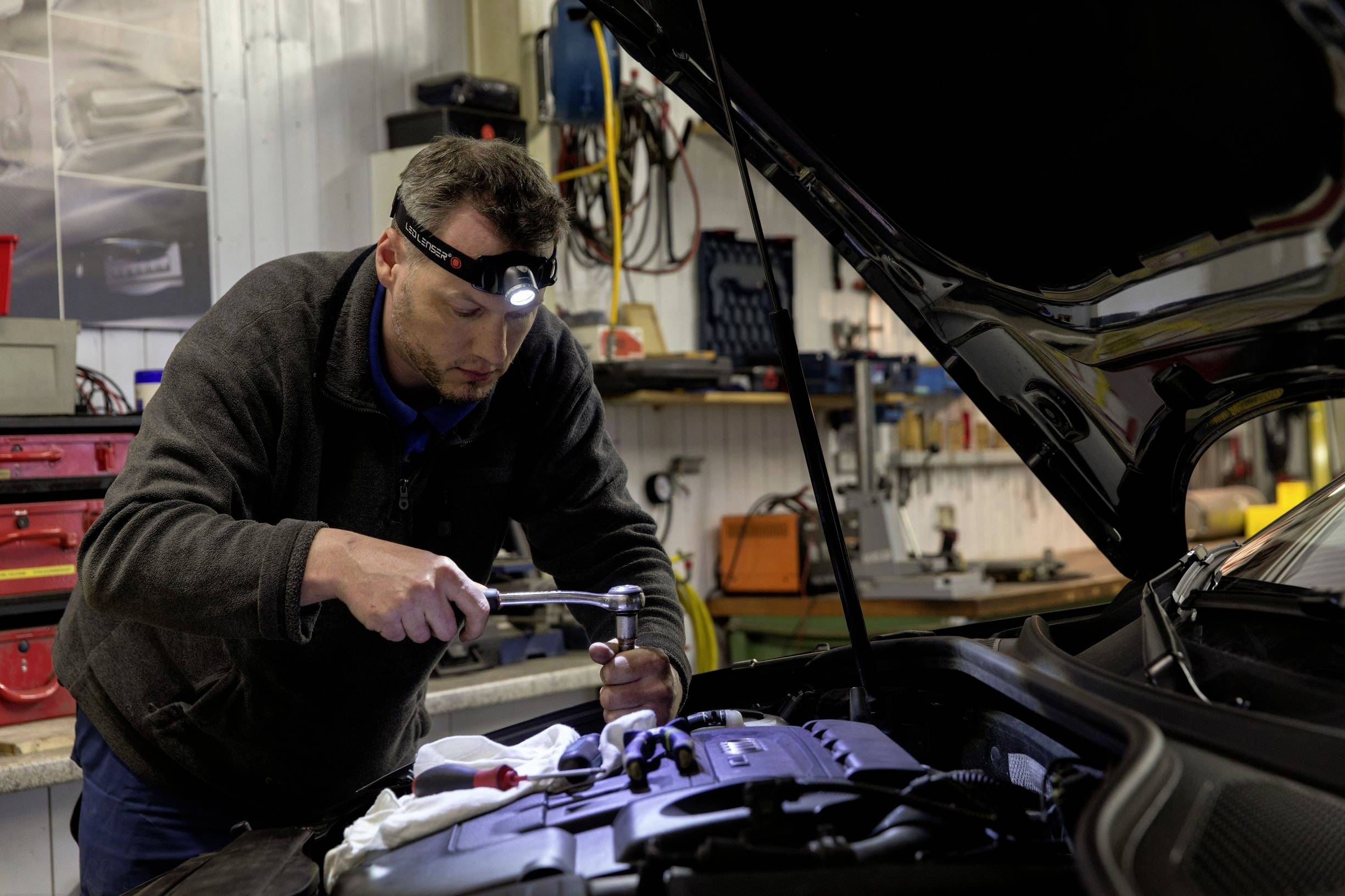 En mekaniker arbejder med en lommelygte ved den åbne motor på en bil i et værksted. Værktøj og udstyr er synlige i baggrunden.