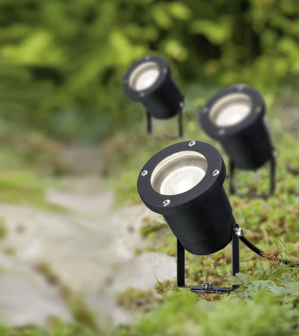 Tre sorte jordrettede spotlights er installeret på en græsklædt overflade og kaster lys på stien foran dem. I baggrunden ses grøn vegetation.