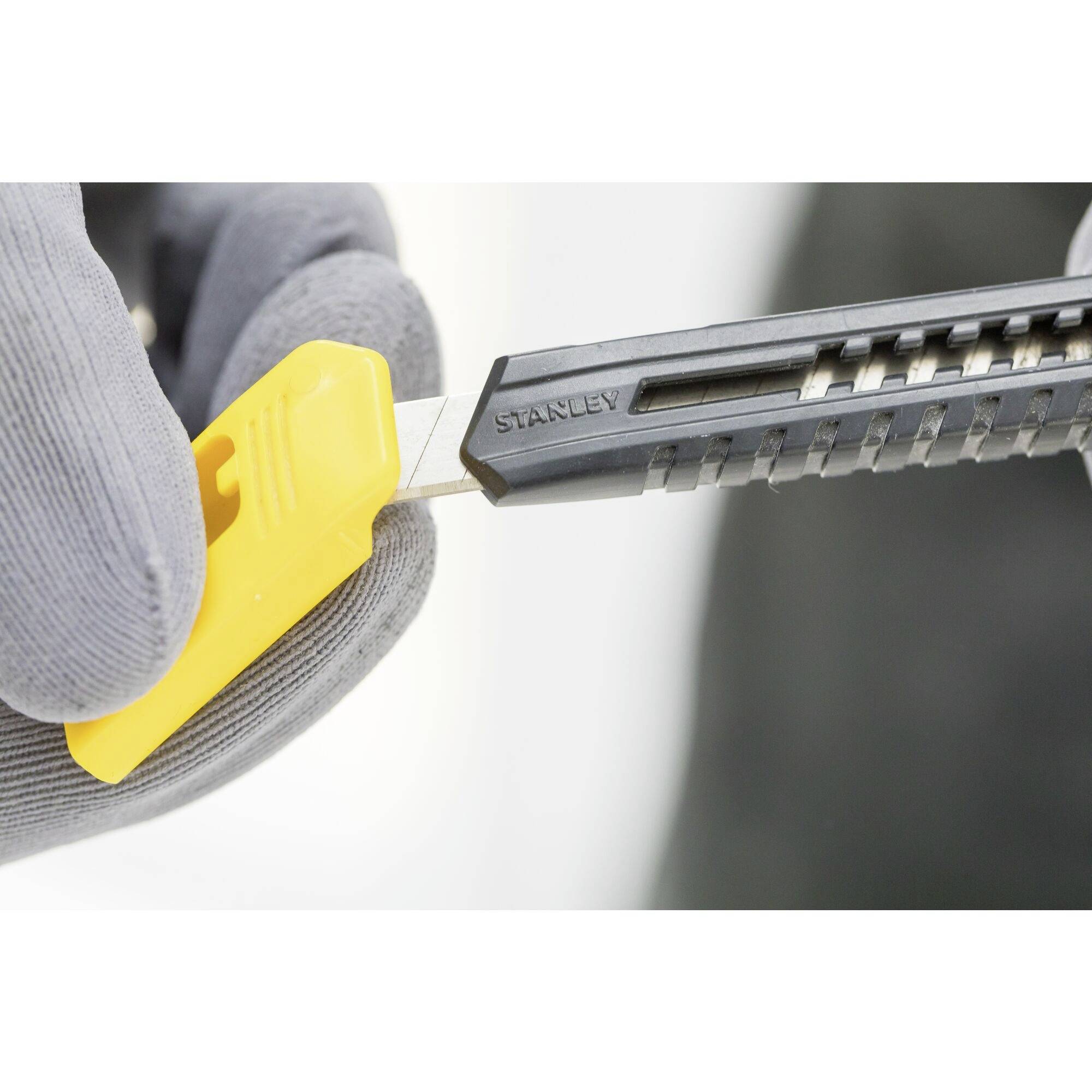 En grå handske holder en gul og sort tæppekniv med en synlig skarp klinge.