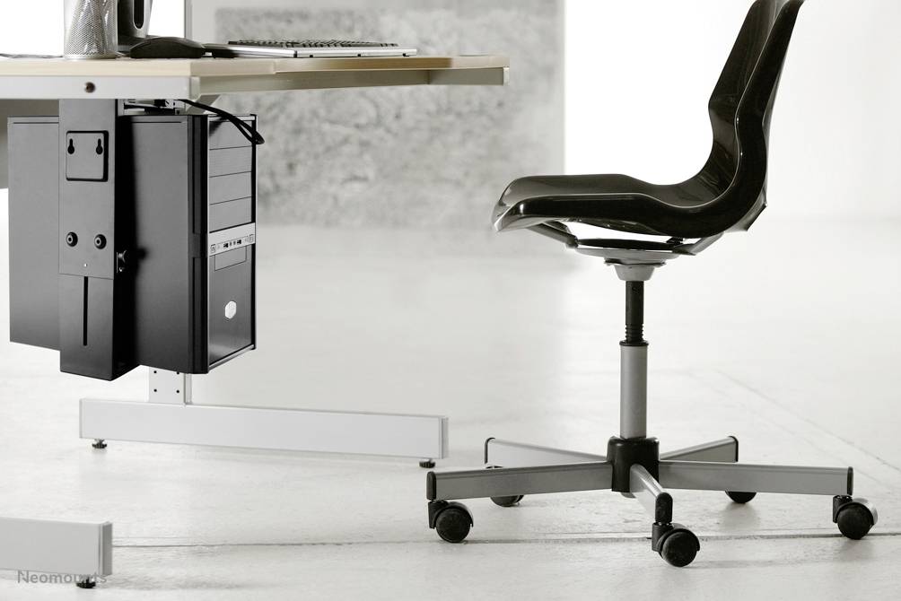 En moderne sort kontorstol med hjul står ved siden af et skrivebord med en computerboks nedenunder, i et minimalistisk rum.