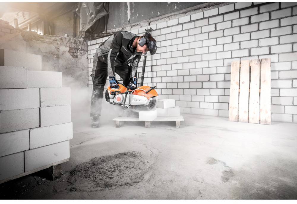 Arbejder skærer betonblokke med en motorsav på en byggeplads. Beskyttelsestøj og støvbeskyttelse er til stede.