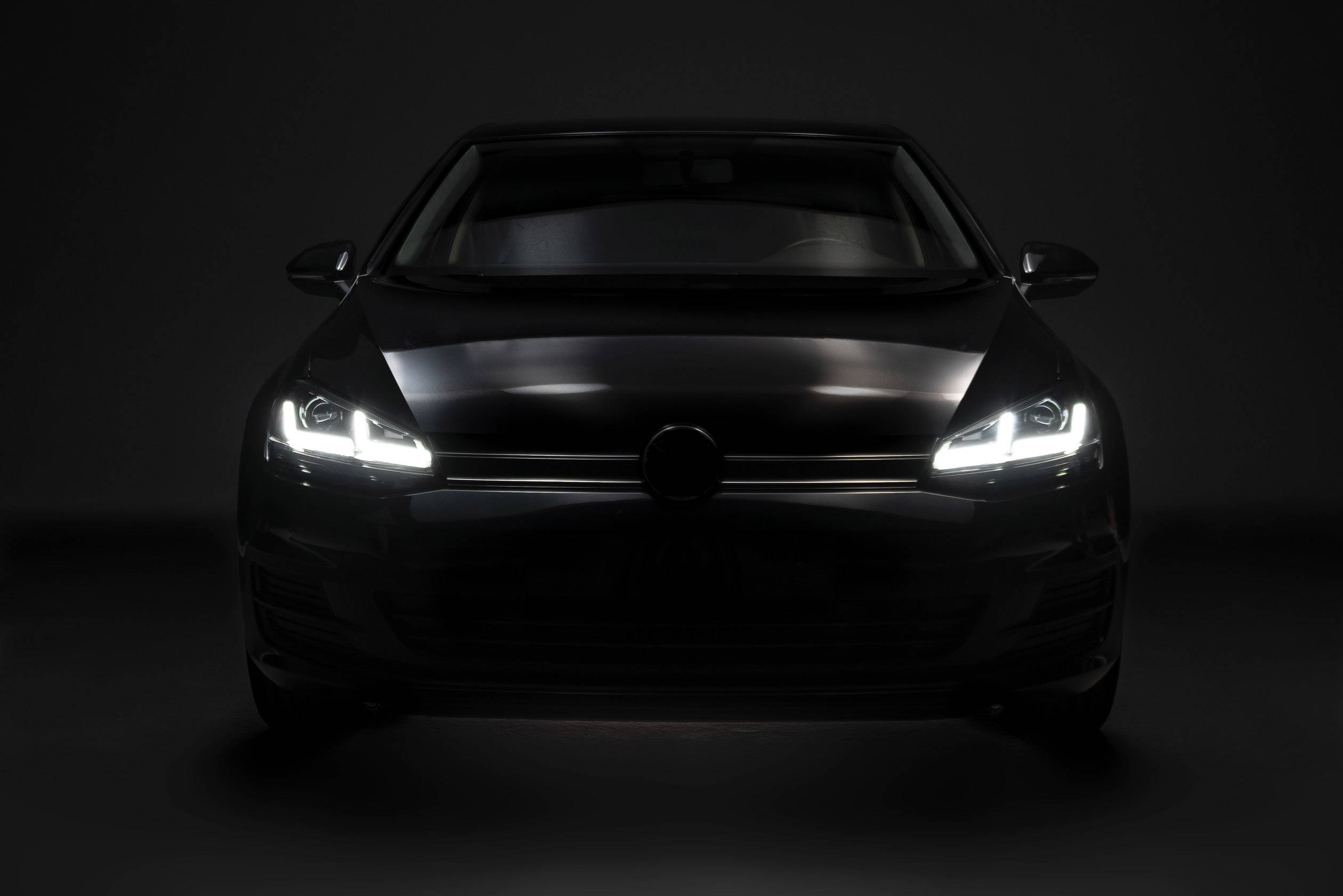En sort bil med tændte forlygter ses forfra, omgivet af en mørk baggrund.