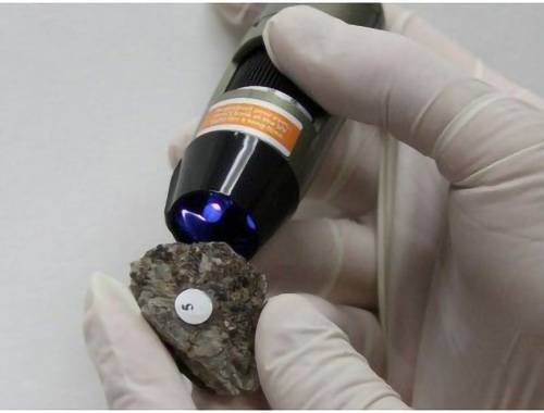 Handsker hænder holder en rockprøve mærket '5', der bliver undersøgt med en lysudslippende videnskabelig enhed.