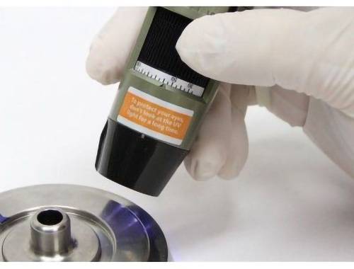 En handskeklædt hånd påfører lim fra en tube på en metaloverflade, med tekst på tuben, der advarer mod eksponering for UV-lys for effektiv binding.