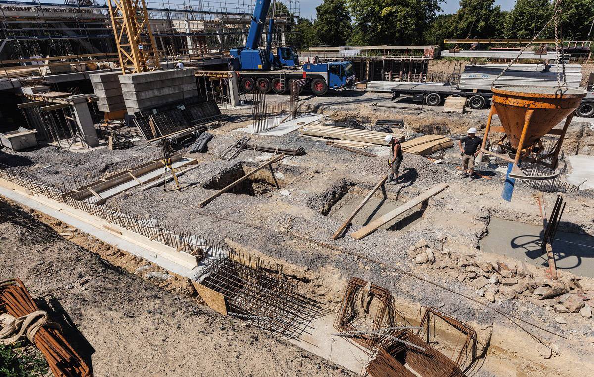 En byggeplads med arbejdere, der er i gang med at støbe et fundament. En kran løfter byggematerialer, og metalstilladser og tunge maskiner er synlige.