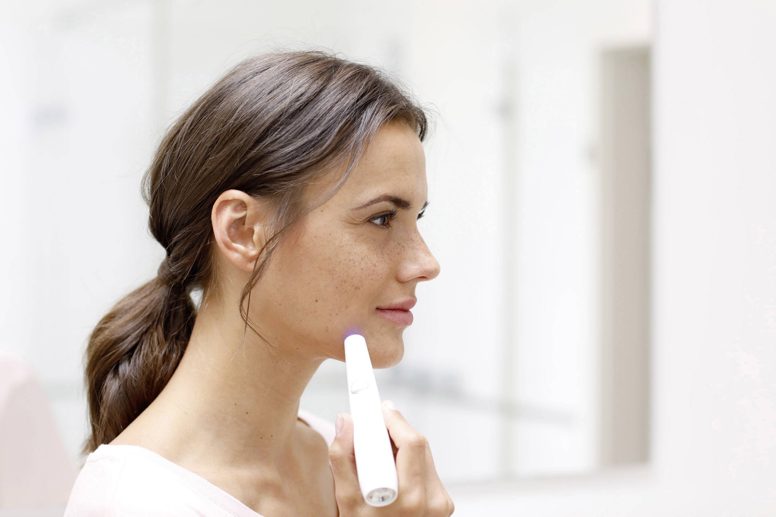 Kvinde med flettet hår holder en hudplejestift op til kinden foran en spejlbaggrund.