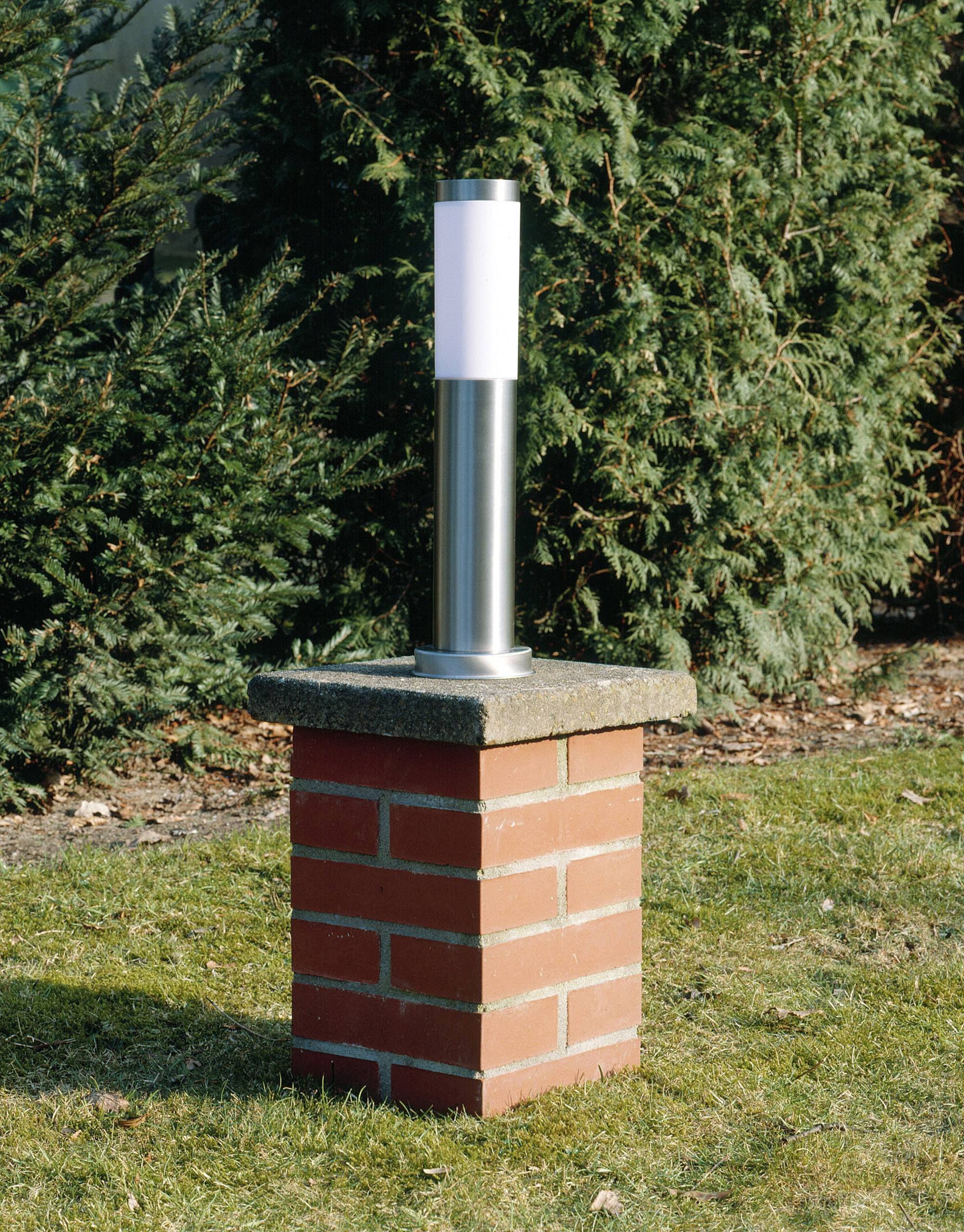 En moderne udendørslampe står på en muret sokkel i en have, omgivet af grønne nålebuske.