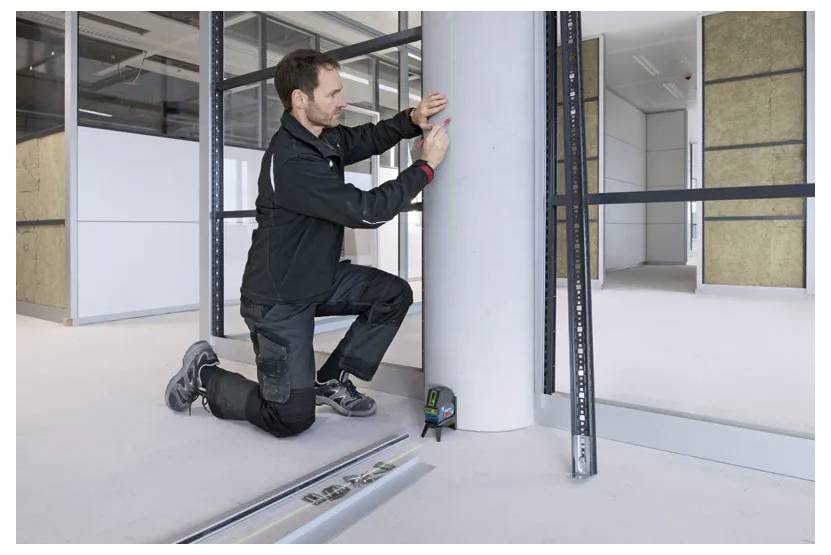 En person knæler, mens vedkommende måler en cylinderformet søjle i et moderne, ufærdigt indvendigt rum med bygningsværktøj i nærheden.