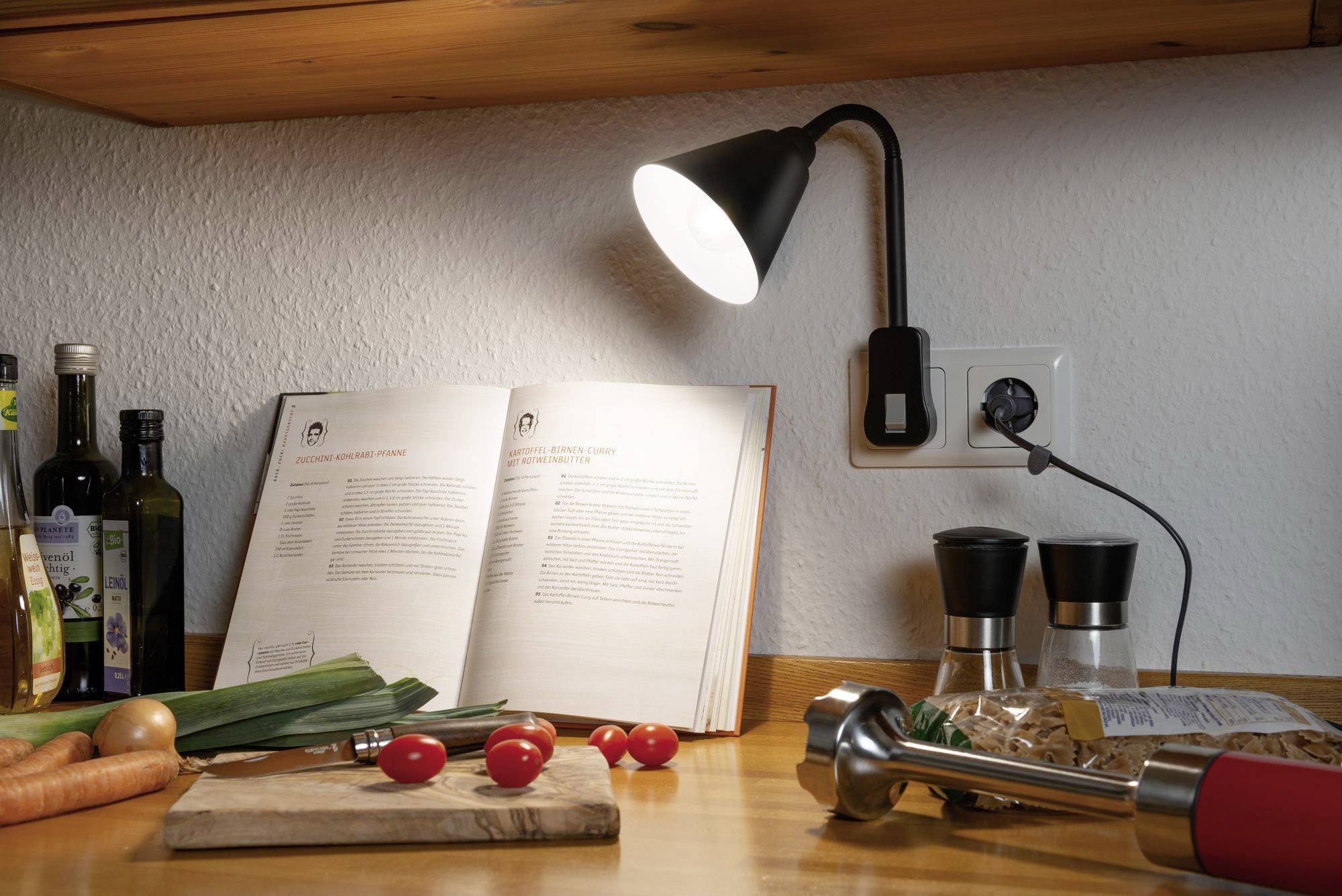 En kogebog ligger åben under en lampe i et køkken. Ved siden af ligger tomater, gulerødder, porre og køkkenredskaber.