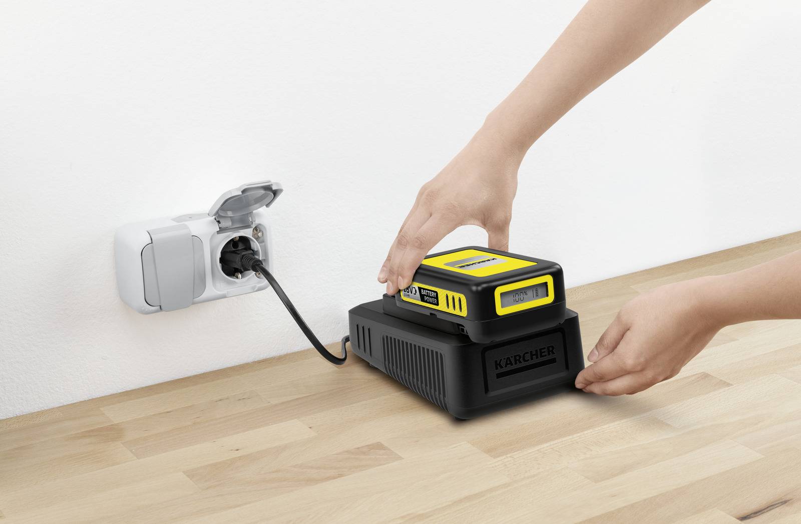 Hænder der placerer et gult og sort batteri i en mærkevarelader på en træoverflade, med en ledning sat i en vægstikdåse.