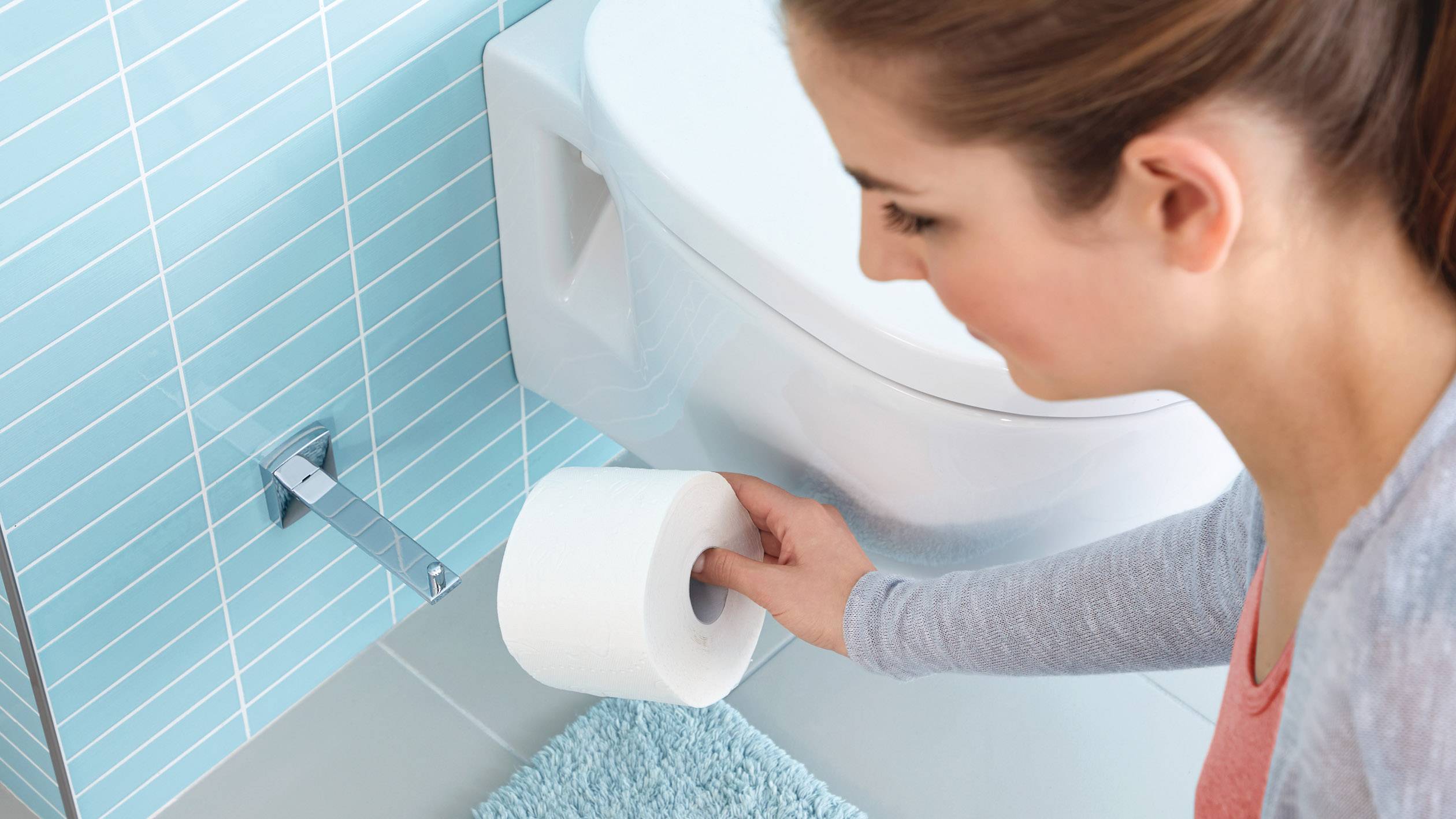 En kvinde rækker efter en rulle toiletpapir i et badeværelse med blå fliser.