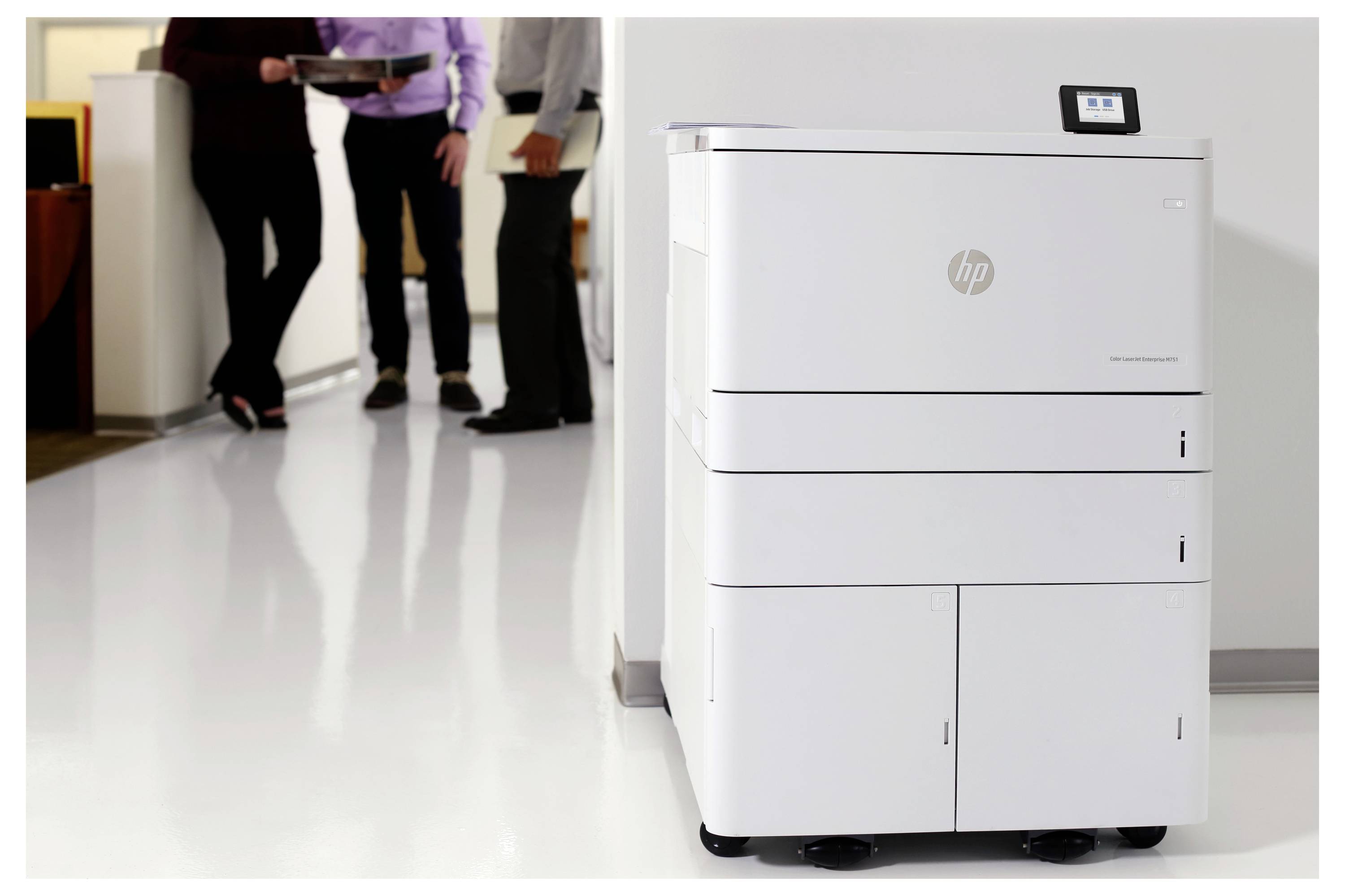 Hvid HP-printer/kopimaskine på en kontorgang med flere personer, der står og taler i baggrunden.