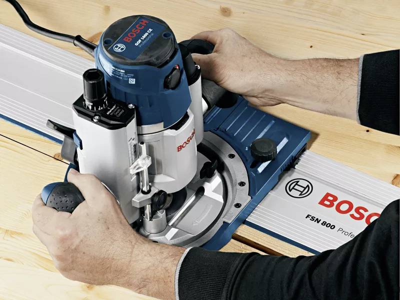 To hænder fører en blå overfræser fra Bosch langs en styreskinner på et træbord.