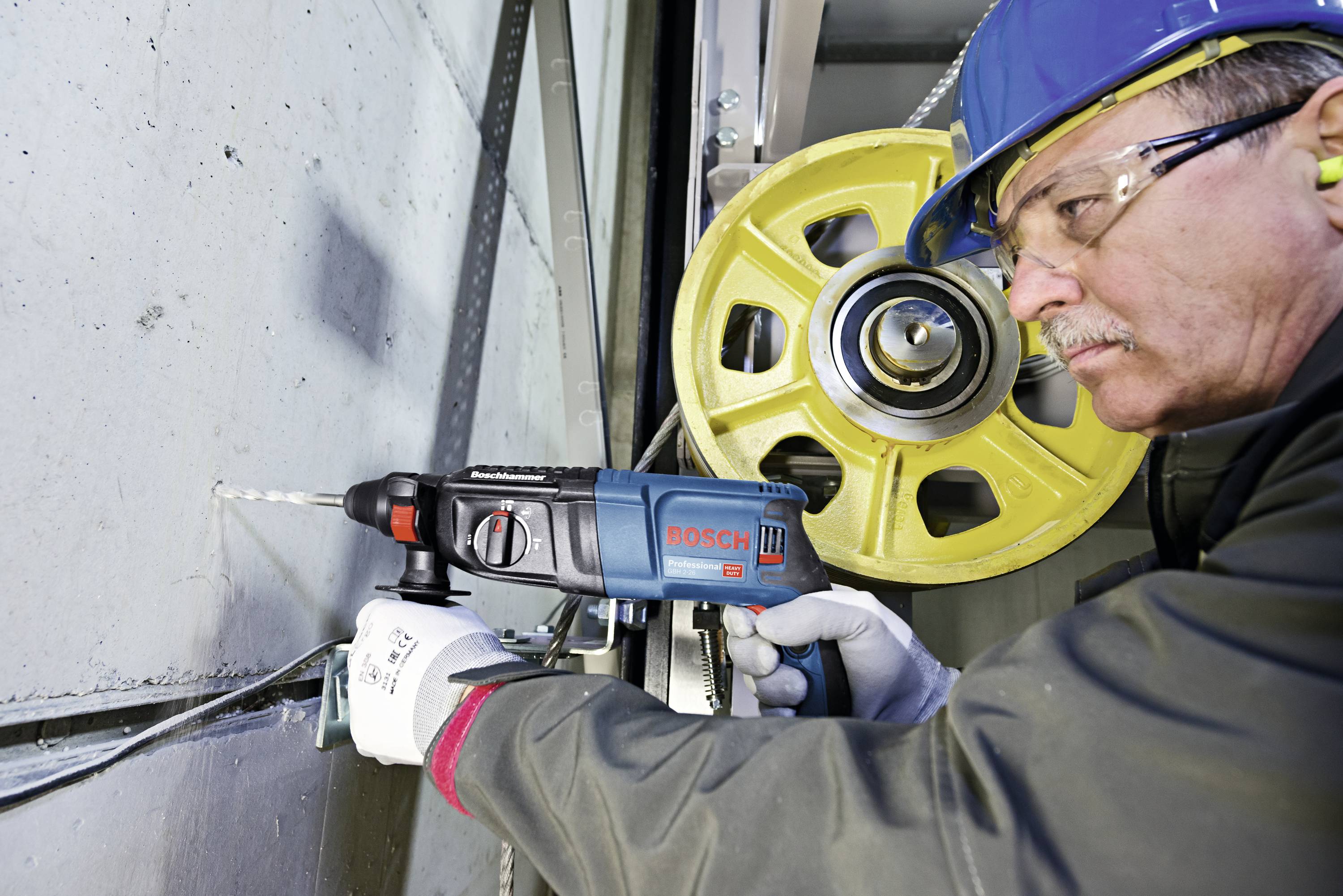En arbejder med sikkerhedshjelm og handsker borer i en væg med en blå Bosch-boremaskine, med en industriel baggrund.