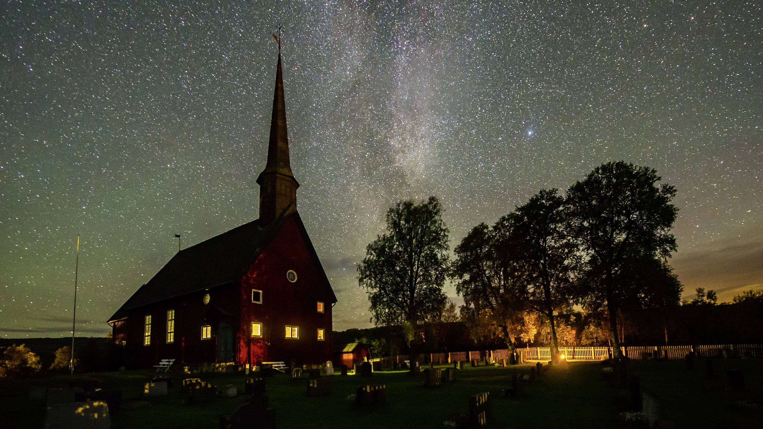 Historisk kirke om natten, belyst af varmt lys, omgivet af træer. Den klare stjernehimmel er synlig i baggrunden.