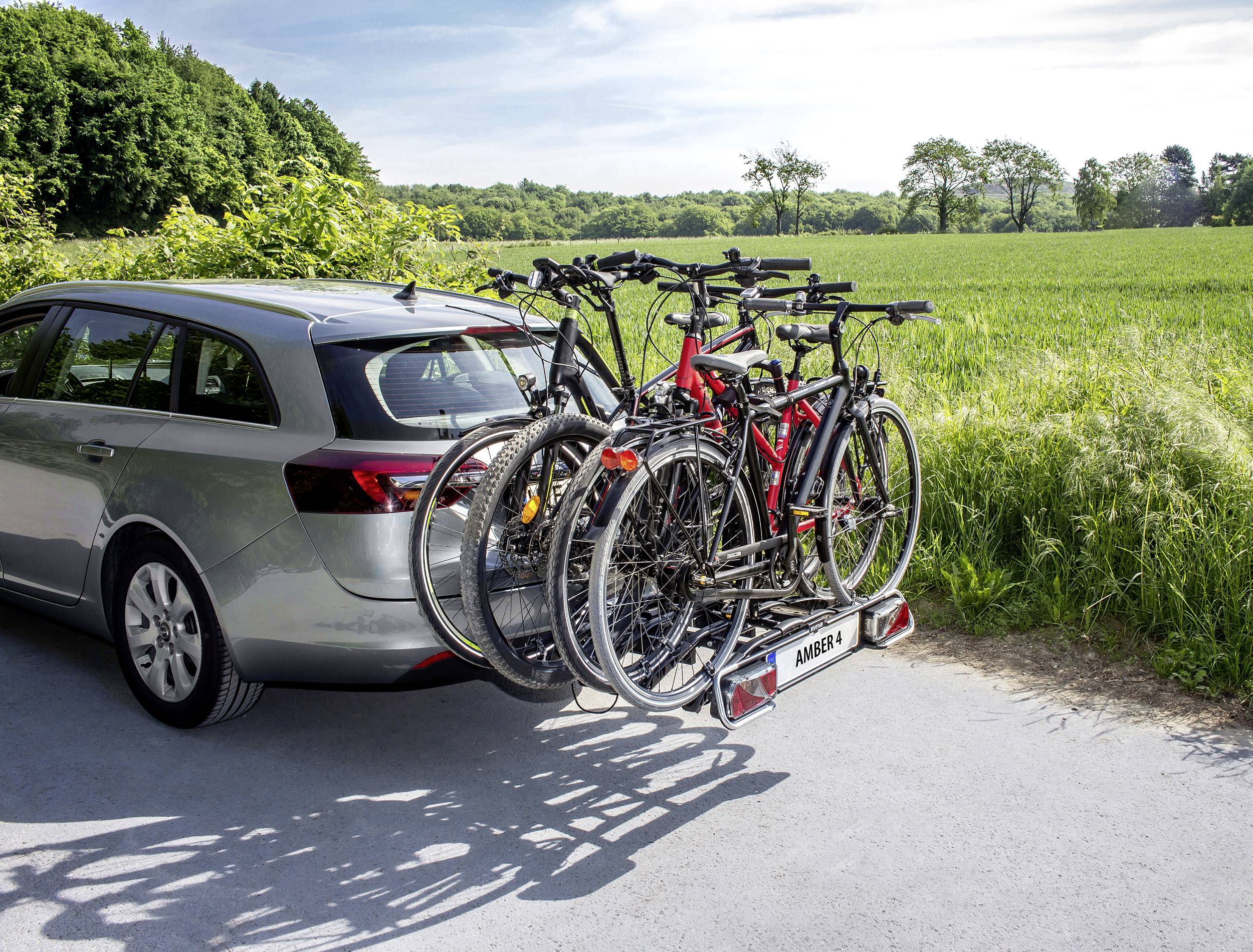 En sølvfarvet stationcar kører med fire cykler på en bagmonteret cykelholder. I baggrunden ses en grøn eng og en skov.