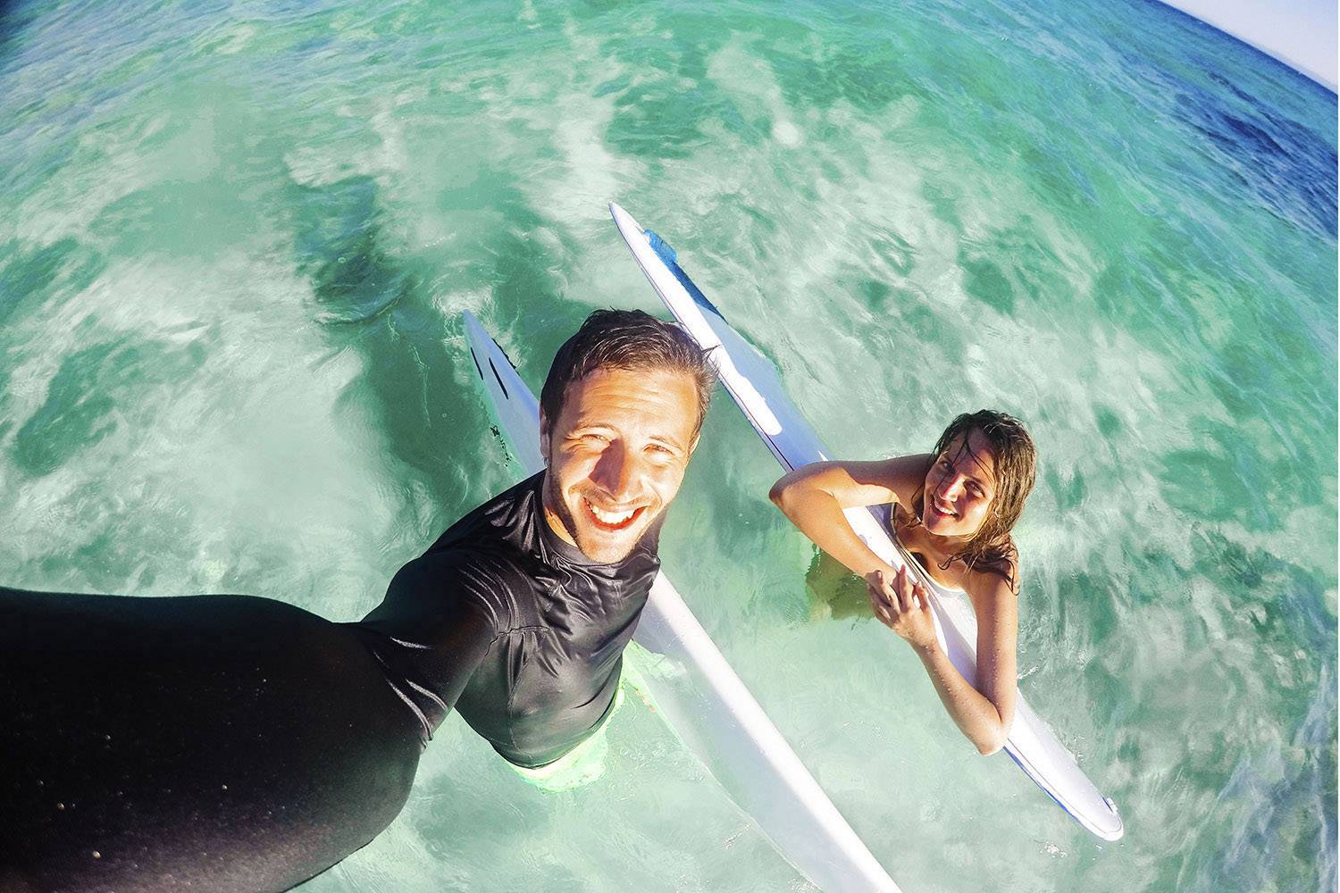 To personer i turkisfarvet vand holder surfbrædder og smiler til kameraet på en solrig dag med klar himmel.