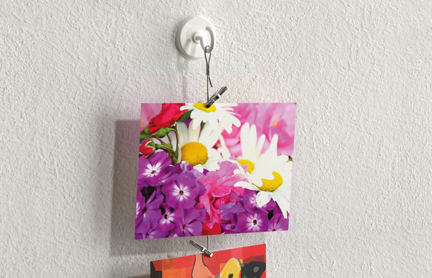 Et blomsterfototryk af tusindfryd og lilla blomster fastklippet på en vægkrog med et lille metalclip, med et andet tryk delvist synligt nedenfor.