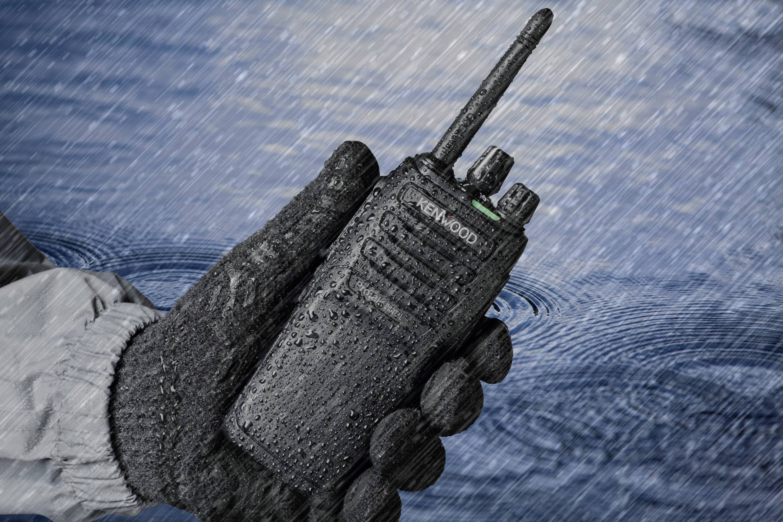 Et vandbelagt radioapparat holdes af en handsket hånd i regnen, hvilket viser dets robusthed og vandtæthed.