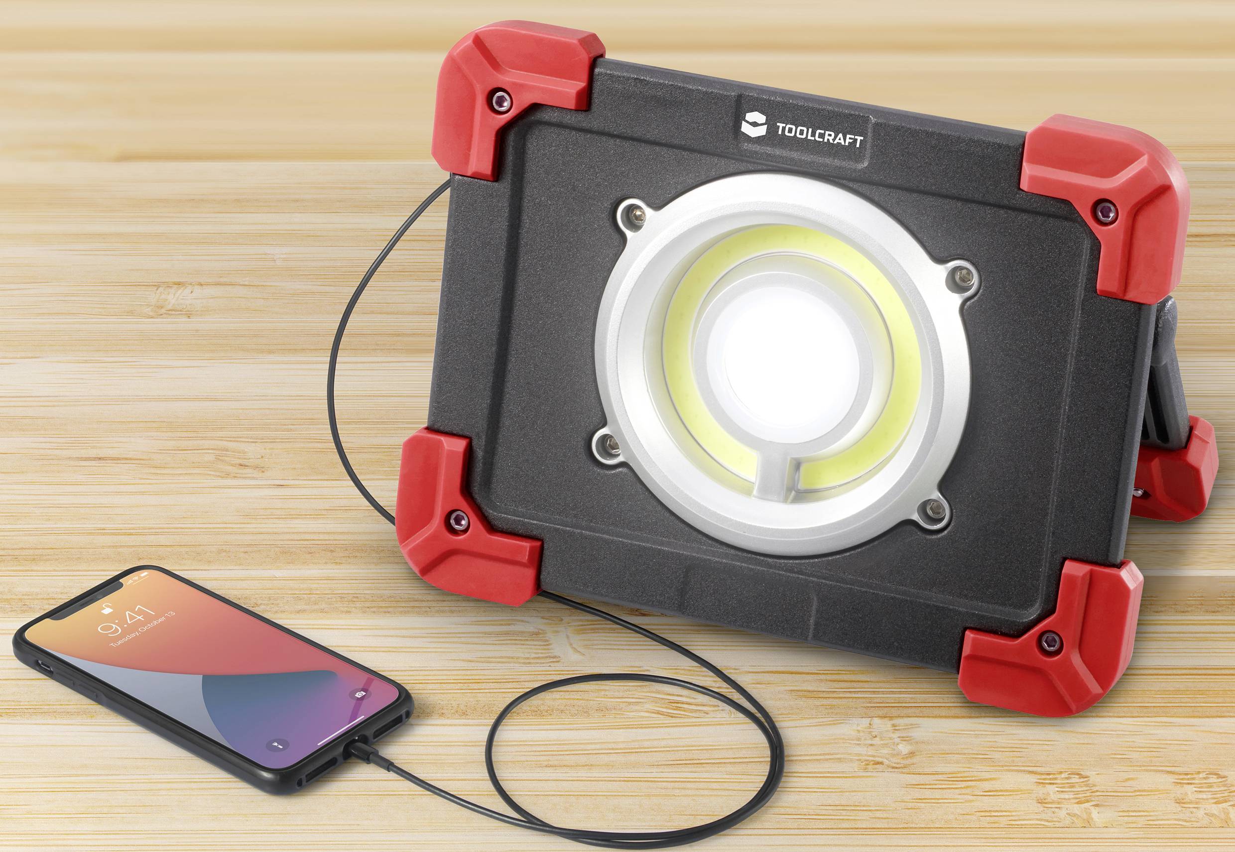 Bærbar LED-arbejdslampe med rød ramme oplader en smartphone via USB-kabel. Den står på et træbord.