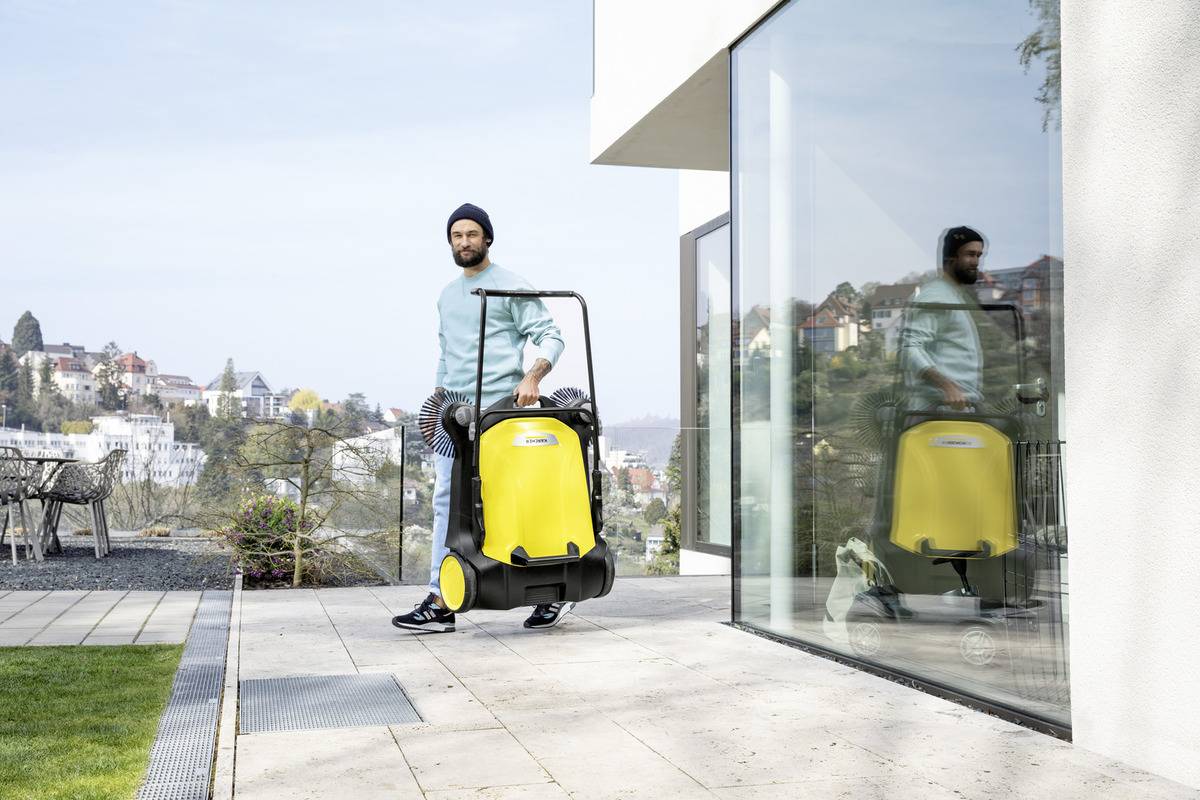En person står uden for en moderne bygning og holder en gul rengøringsmaskine. Beboelsesområde med huse synlige i baggrunden.