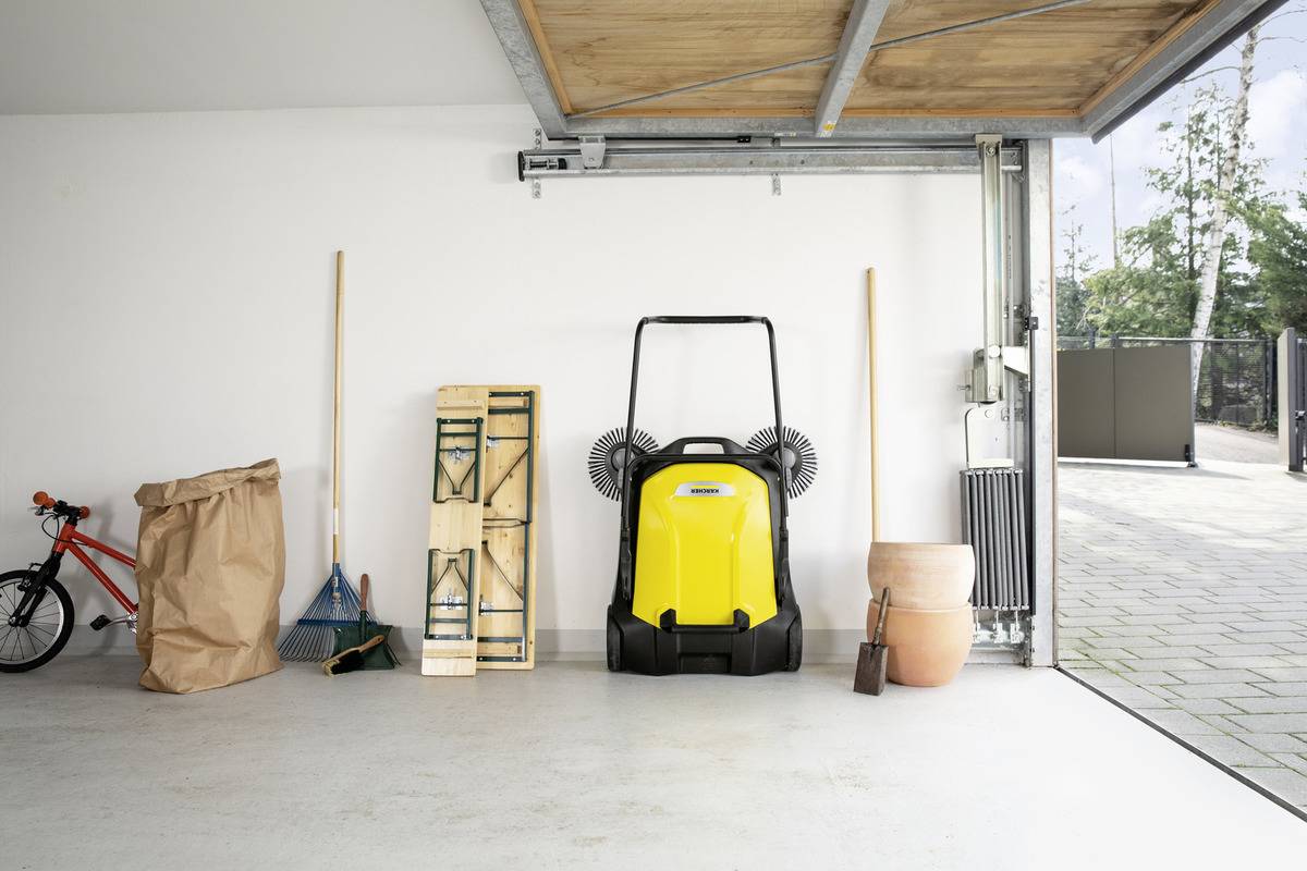 En velorganiseret garage med en gul gulvfejer, værktøj og en cykel pænt placeret op ad væggen; den åbne garagedør viser indkørslen.