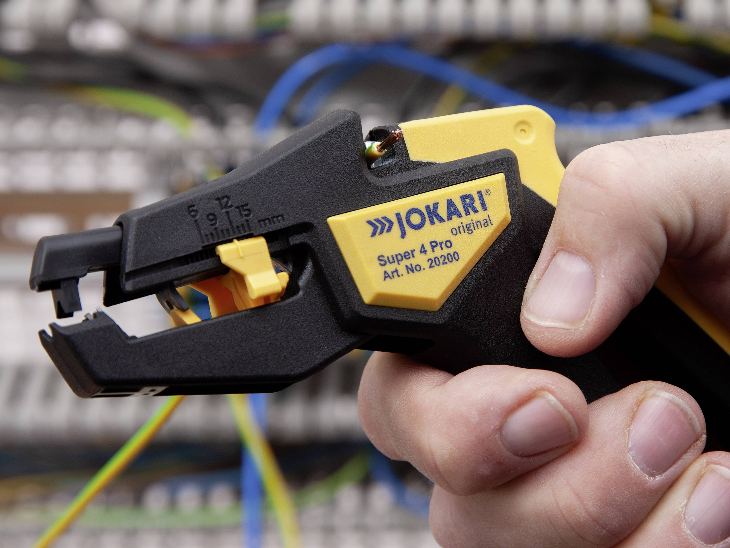 En hånd holder en Jokari Super 4 Pro afisoleringsværktøj foran en baggrund med elektriske kabler.