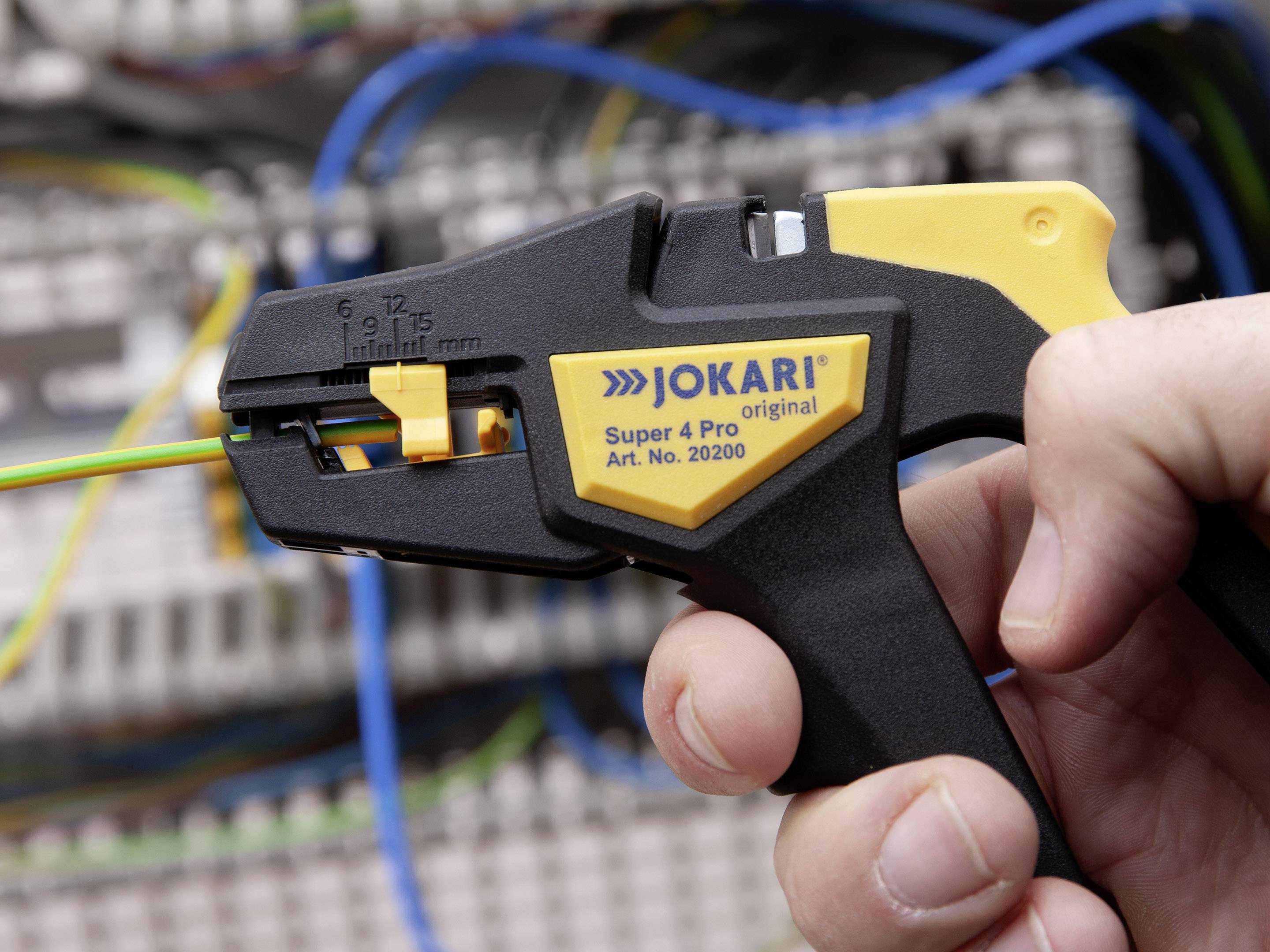 En hånd holder et afisoleringsværktøj foran en uskarp baggrund med elektriske kabler. Værktøjet er velegnet til kabelarbejde.