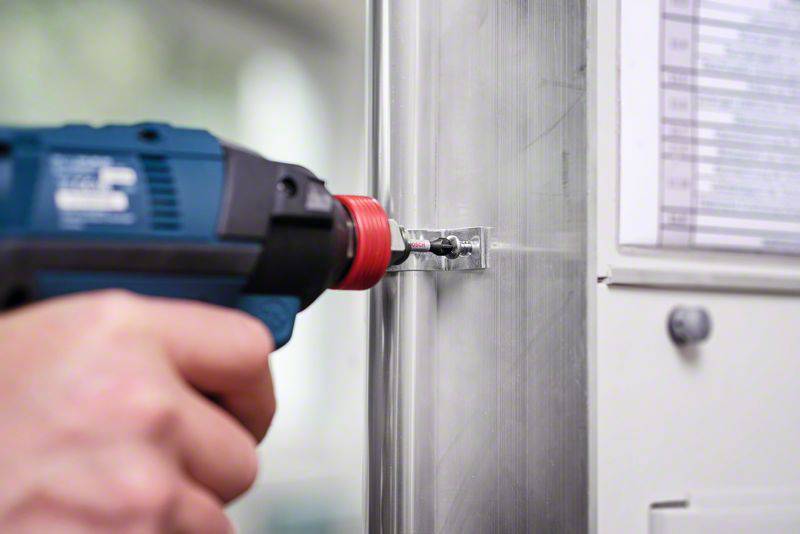 En person holder en blå boremaskine, der borer en skrue i en metaloverflade. Fokus er på værktøjets præcision.