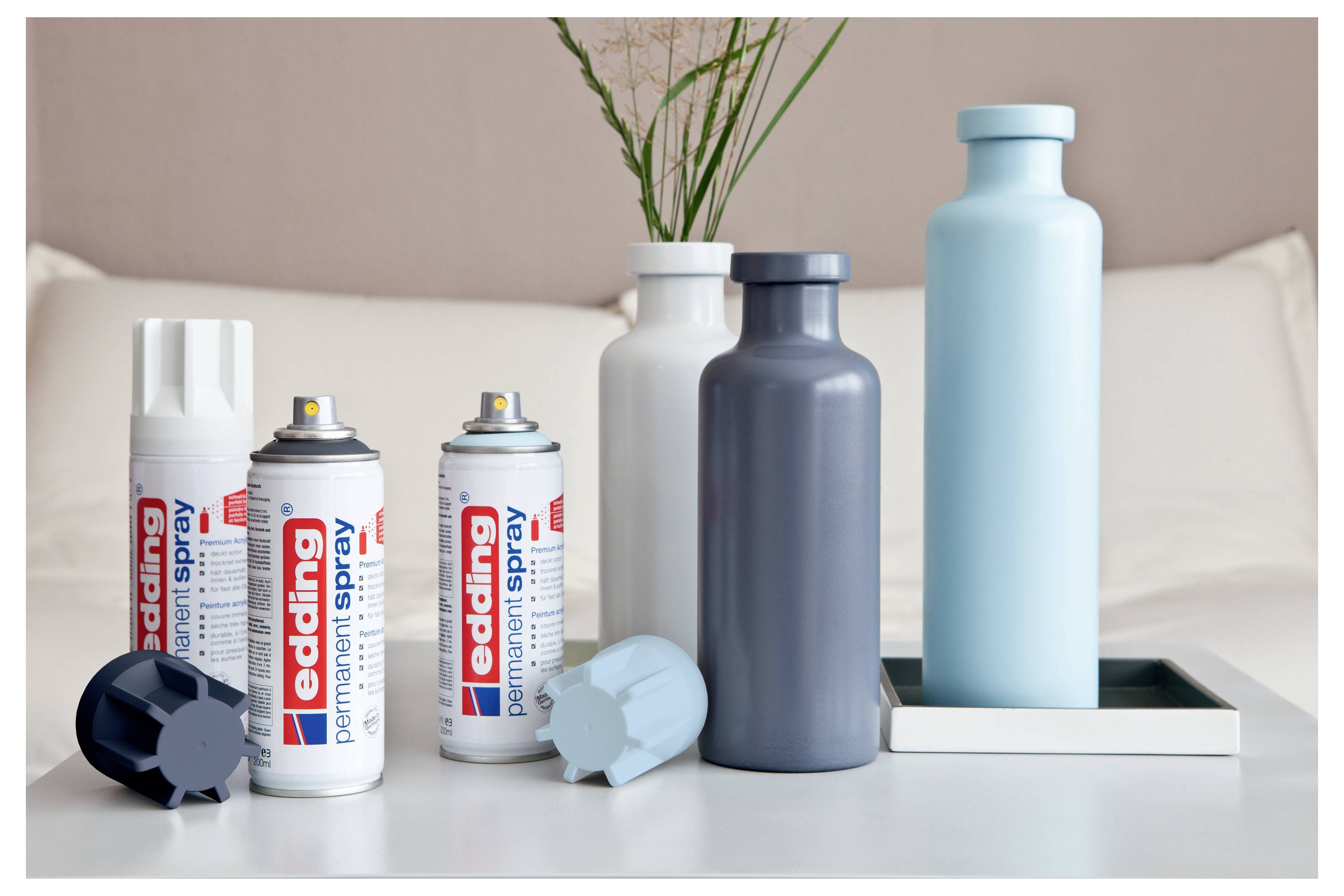 Diverse sprayflasker og genanvendelige flasker arrangeret som et produktdisplay på et bord, med neutral hjemmeindretning i baggrunden.