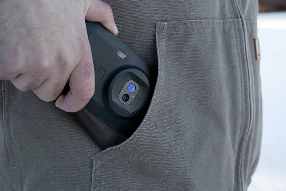 En person stikker en mobiltelefon i en jakkelomme. Mobiltelefonen har en fremhævet blå tast og et sort cover.