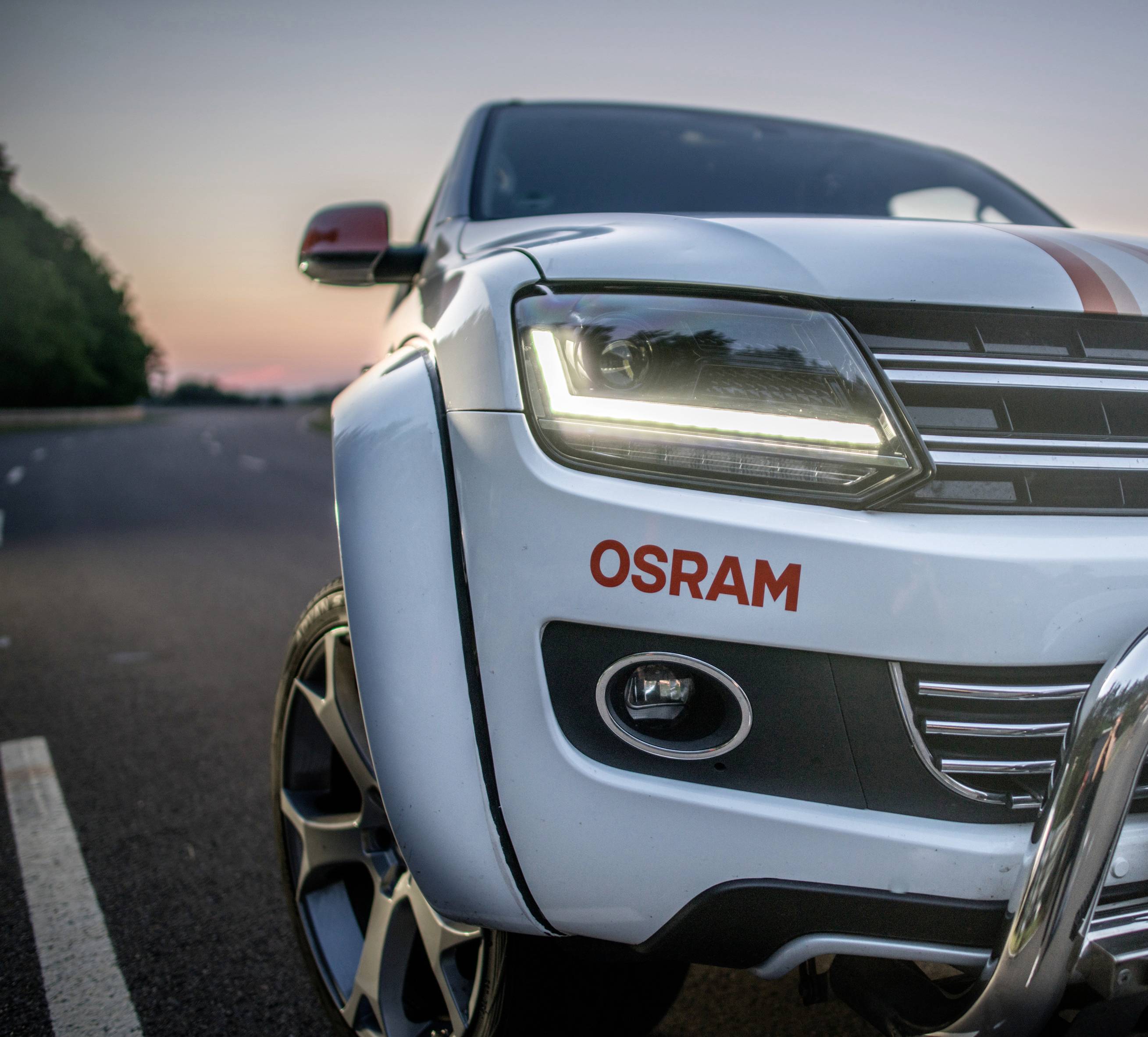 Hvid bil med OSRAM-logo fortil på landevej ved solnedgang, nærlys tændt, vejen drejer til venstre.