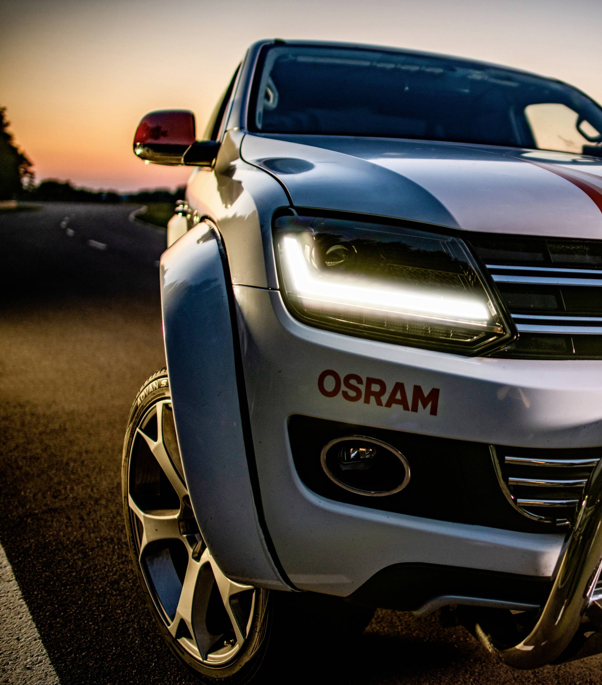 En hvid bil med tændte forlygter holder på en vej ved solnedgang, 'OSRAM' er angivet på fronten.
