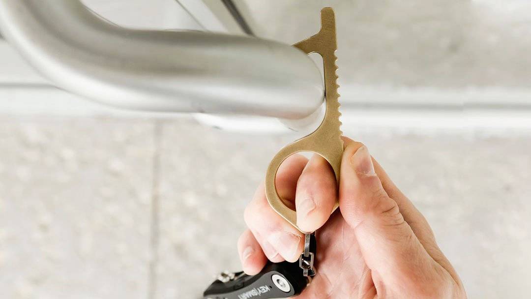 En hånd bruger et berøringsfrit metalværktøj til at åbne en dør og derved undgå direkte kontakt med dørhåndtaget.