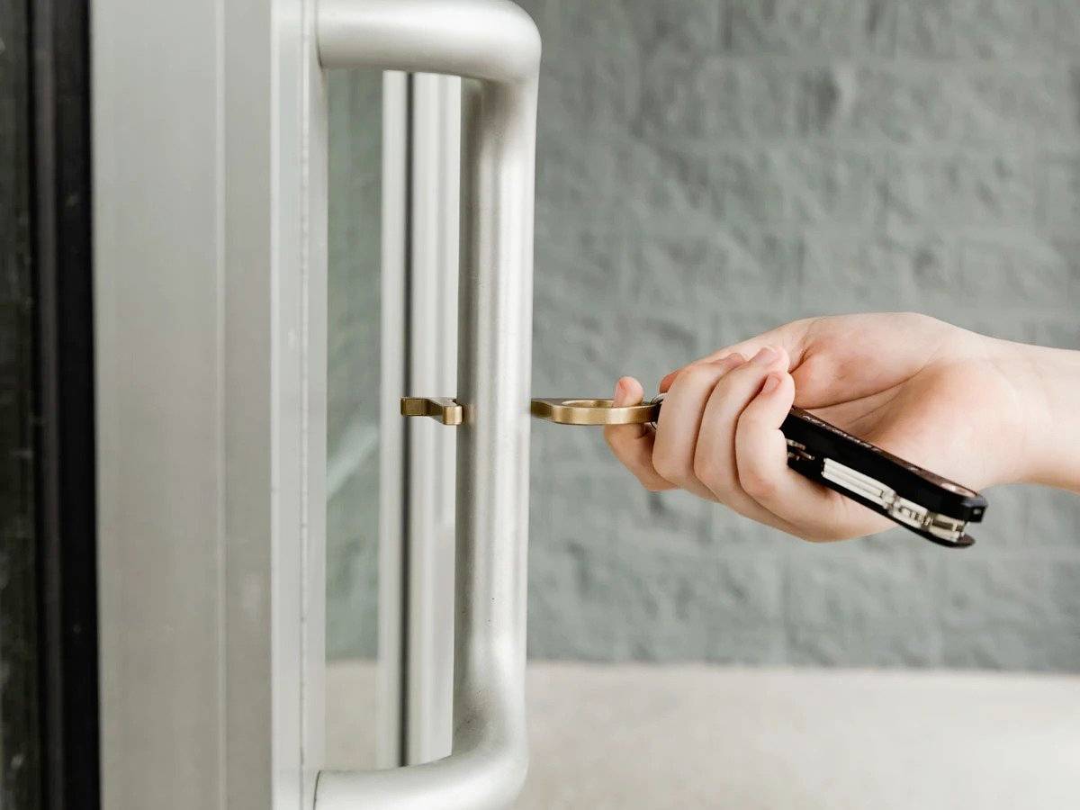 En person åbner en glasdør med en nøgle, holder også en sort foldekniv. Dørammen og den grå væg er synlige.
