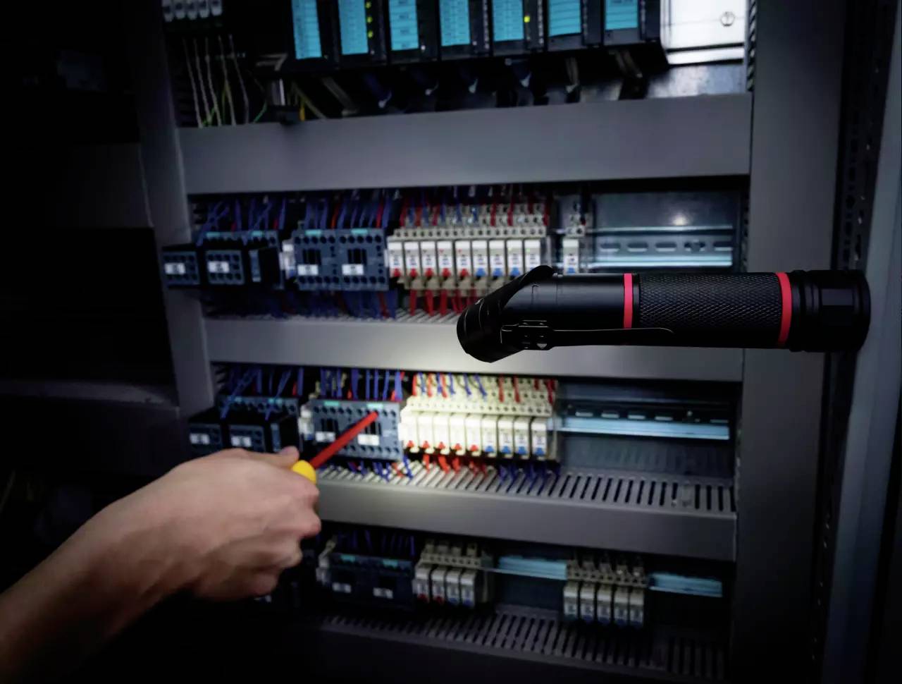 Hånd peger med en skruetrækker på et elektrisk anlæg, belyst af en lommelygte. Teknisk og vedligeholdelsesmæssig kontekst.