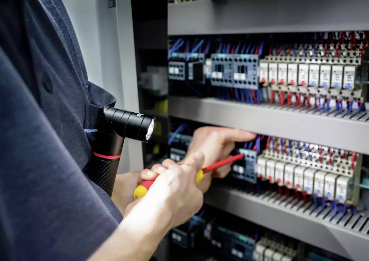 En person arbejder med en skruetrækker på et kontrolpanel fyldt med elektriske komponenter og ledningsføring.