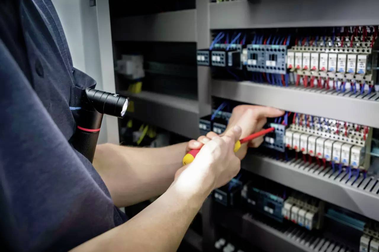 En person arbejder med en skruetrækker ved en åben eltavle fuld af elektriske komponenter og kabler.