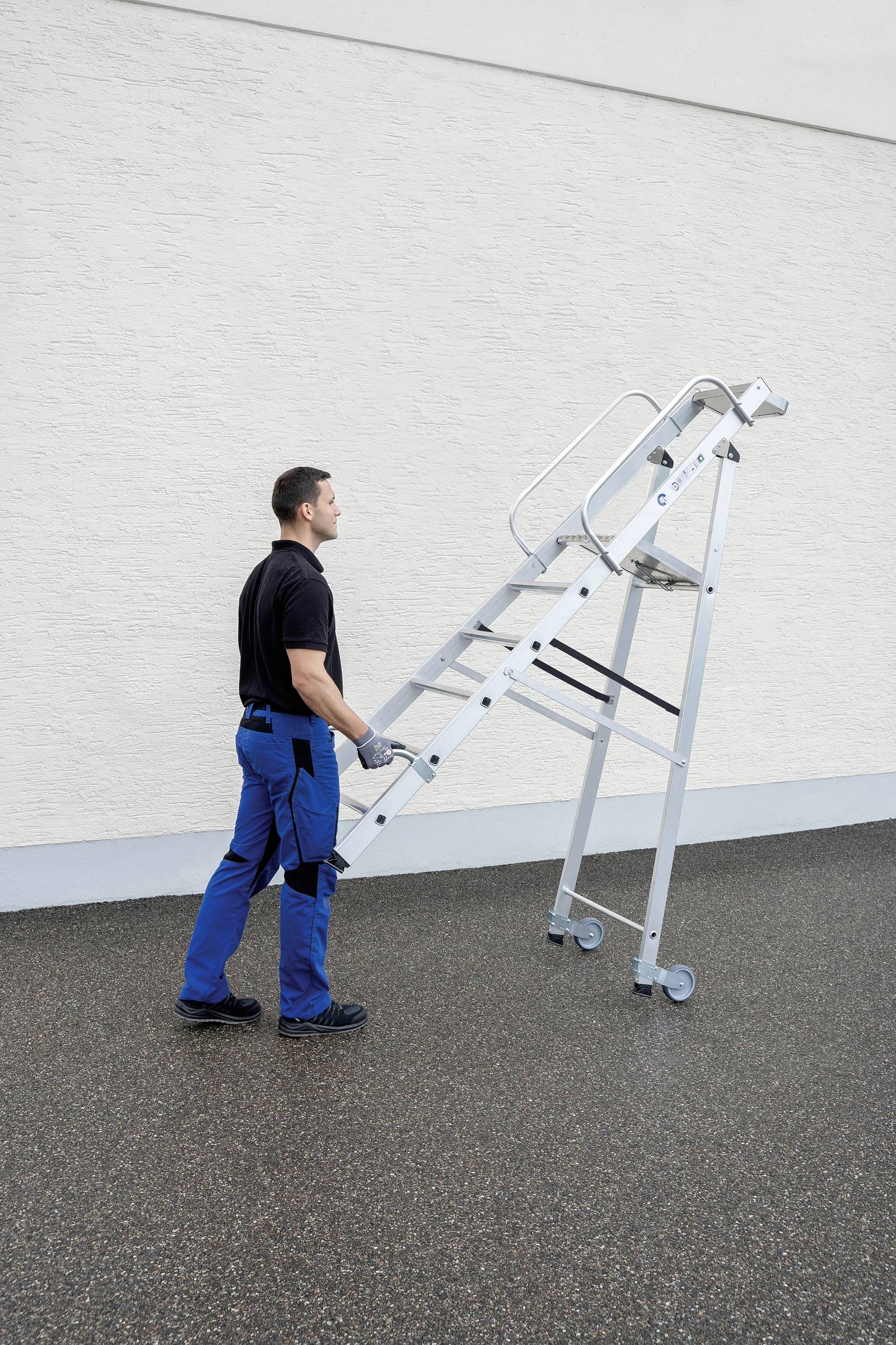En mand i arbejdstøj skubber en mobil aluminiumsstige langs en hvid bygningsfacade på en våd udendørs overflade.