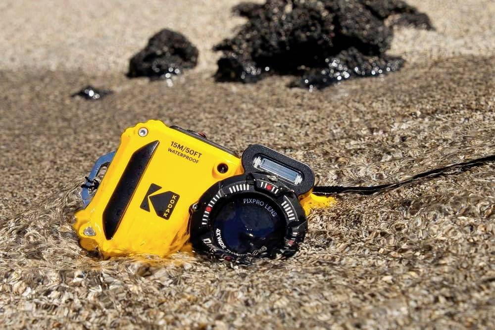 Et gult undervandskamera ligger delvist i vandet på en sandet strand. Sorte klipper er synlige i baggrunden.