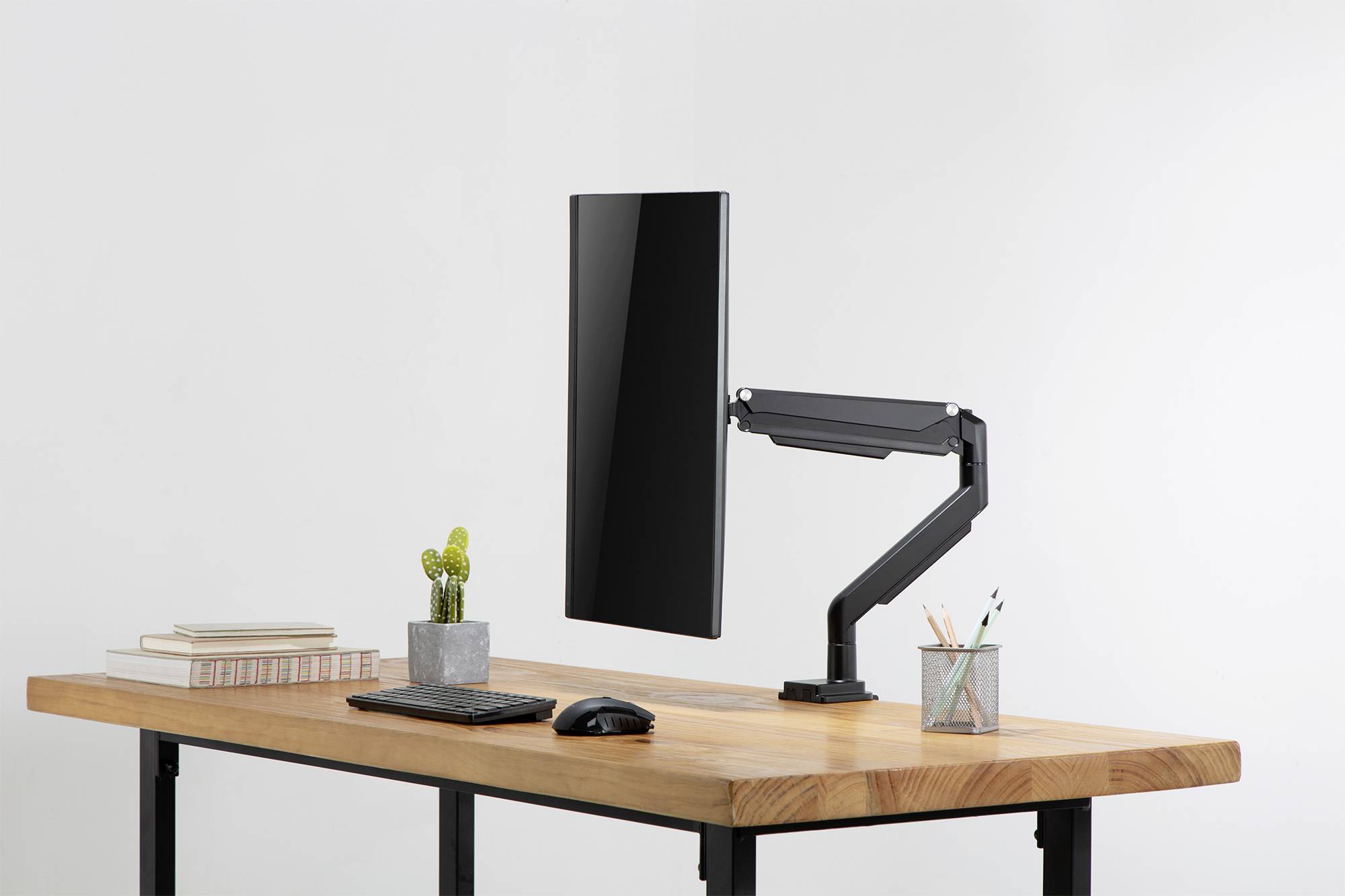 Et moderne skrivebord med en skærm på en svingbar arm, et tastatur, en mus, bøger, en lille kaktus og skriveredskaber.