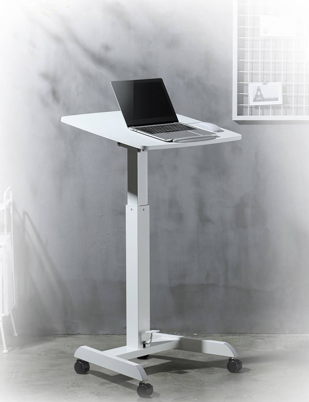Et højdejusterbart hvidt bord med hjul, hvorpå en åben bærbar computer står, foran en grå væg og et hvidt tremågitter.