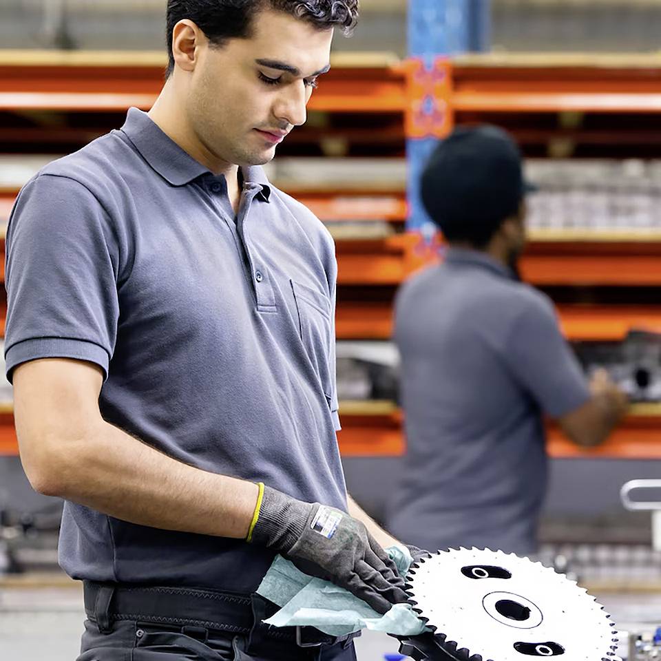 En person i et værksted inspicerer et tandhjul, iført sikkerhedshandsker og en poloskjorte. I baggrunden er hylder fyldt med værktøj og udstyr.