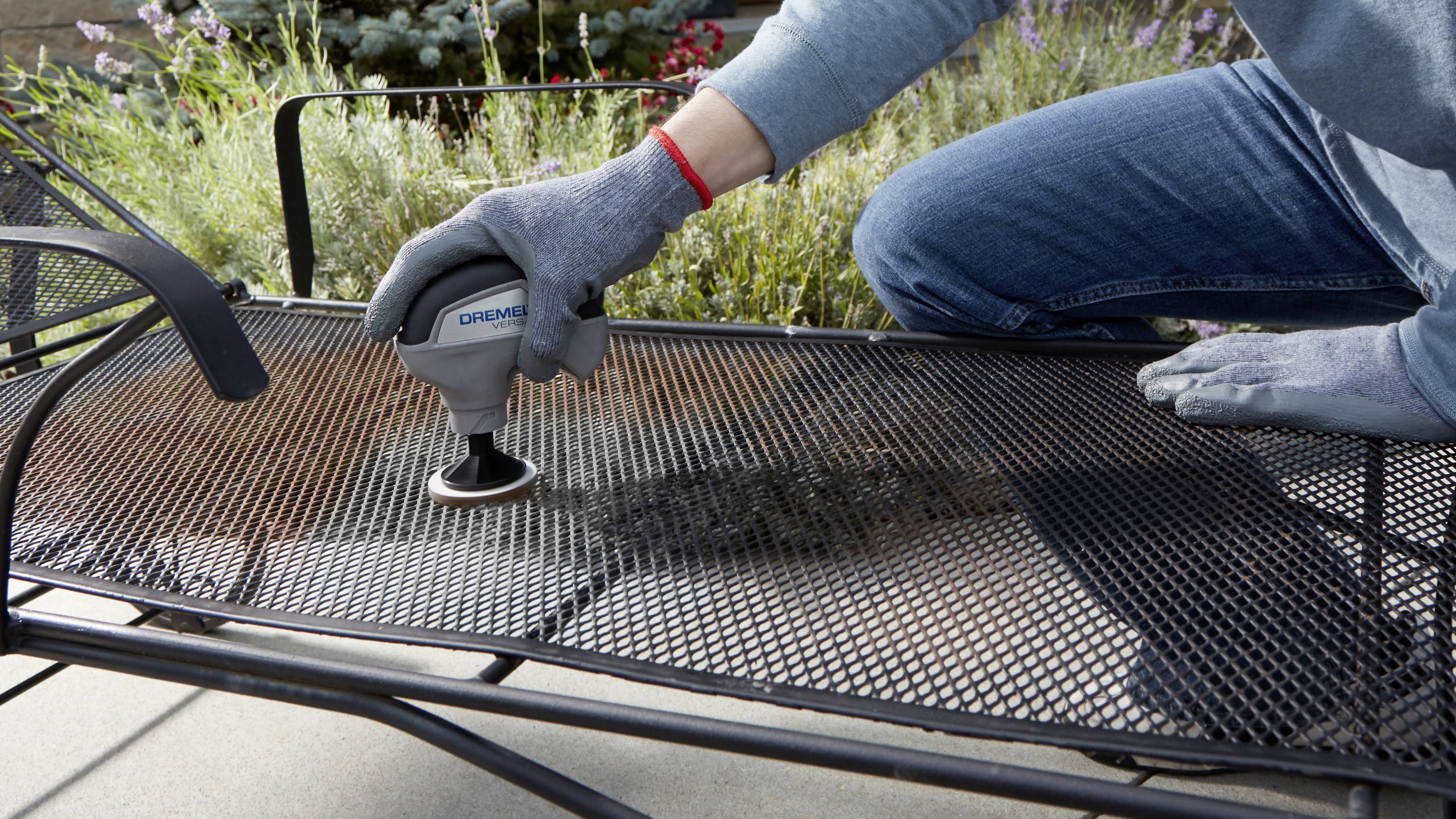 En person rengør et metaltrådsmøbel udendørs med et elektrisk værktøj. I baggrunden ses planter.