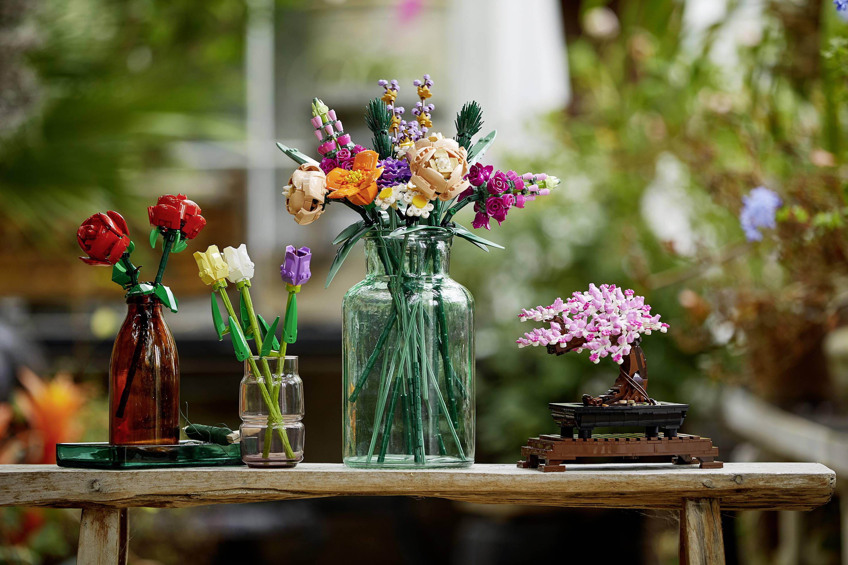 Flere blomsterbuketter i glas på et træbord, herunder en lille bonsai. I baggrunden ses slørede planter.