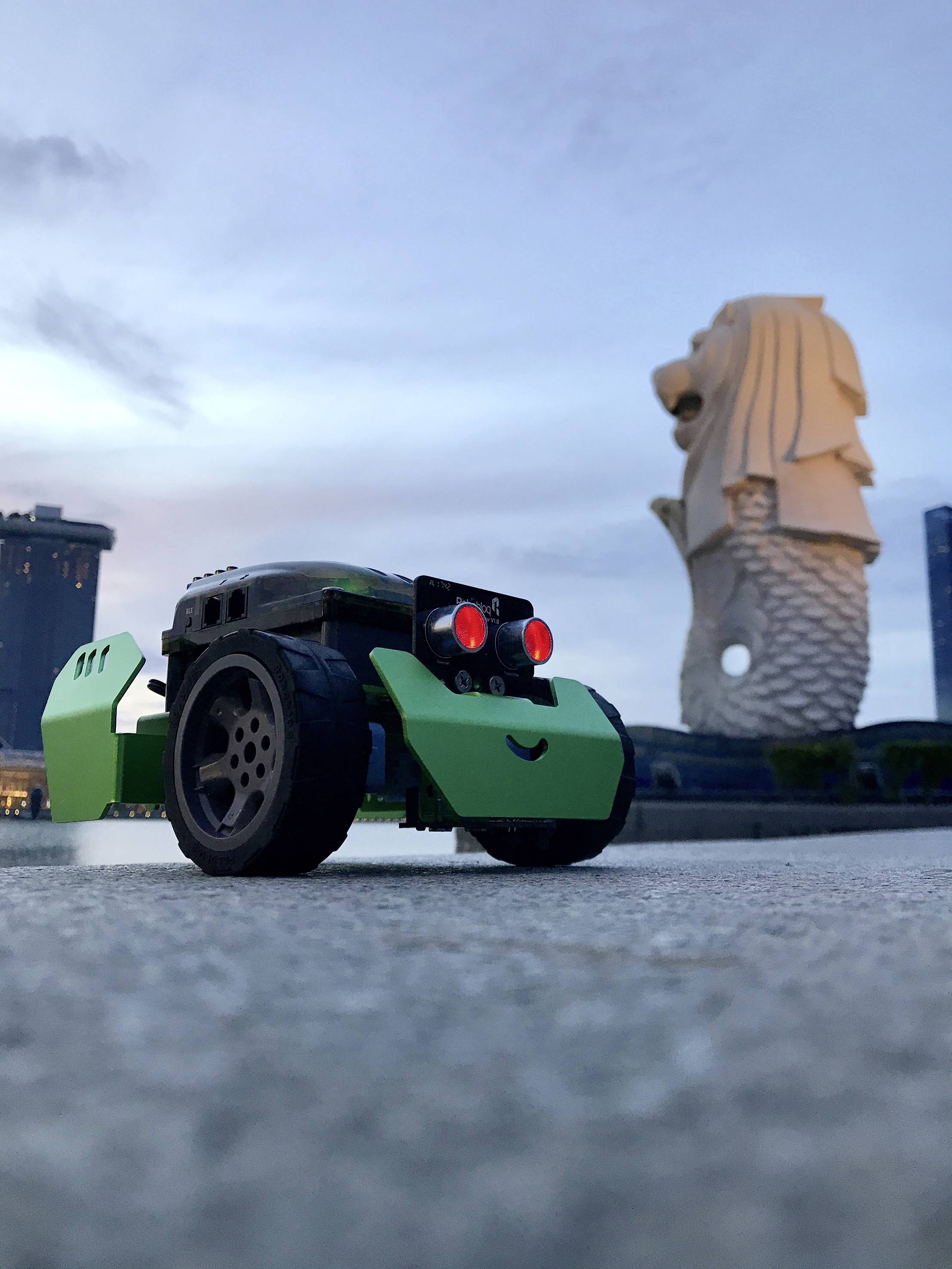 Et lille robotkøretøj med rødt lysende øjne står på et stengulv. I baggrunden ses Merlion-statuen.