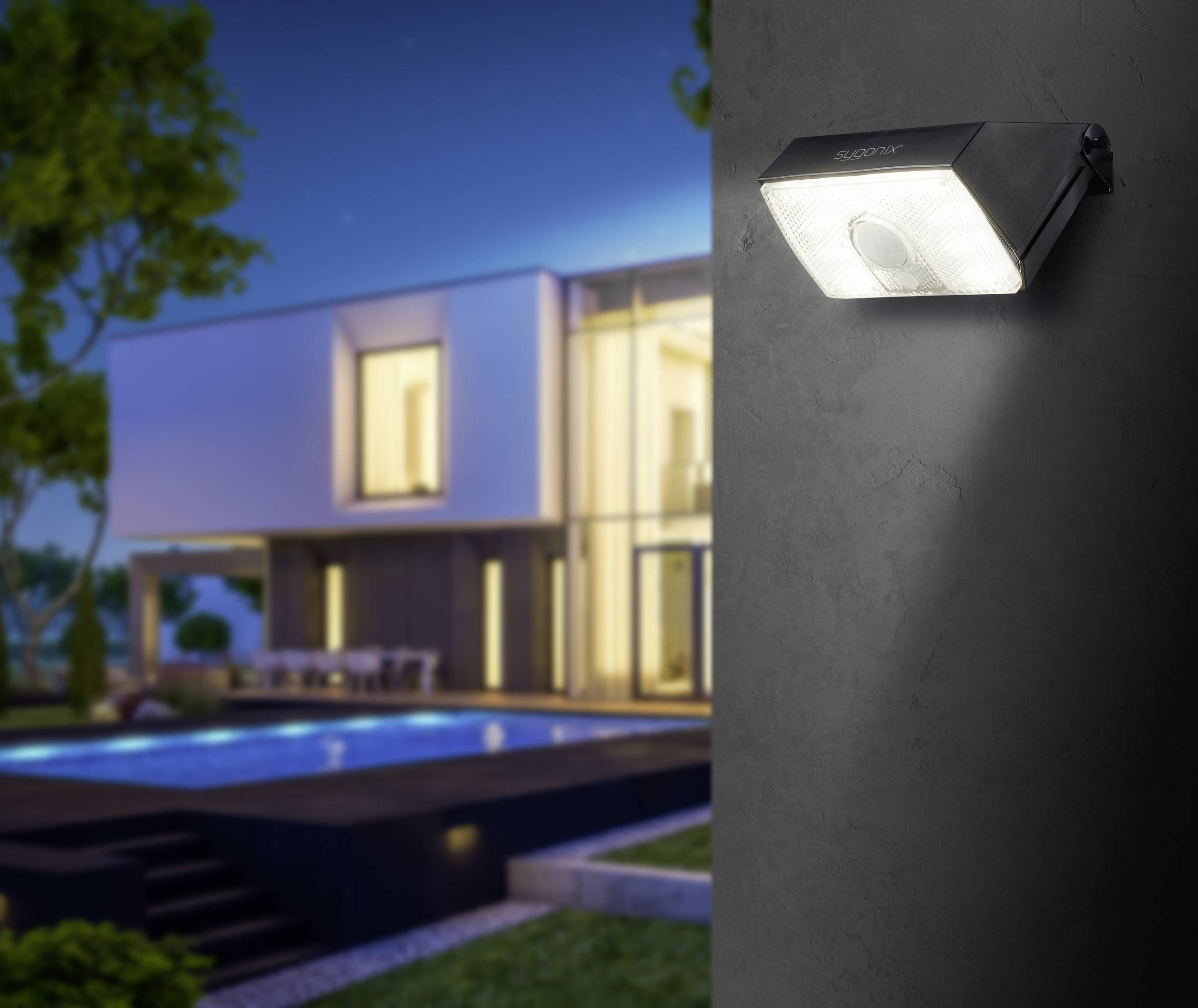 Udvendig vue af et moderne hus om natten, belyst af en væglampe i forgrunden. En swimmingpool er synlig i baggrunden.